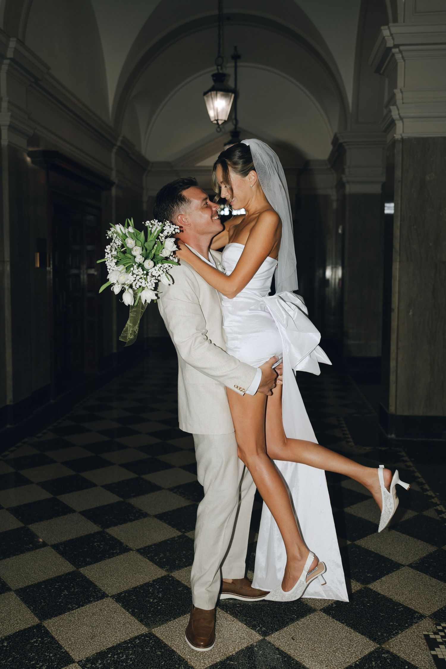 A romantic wedding couple dances and embraces in an elegant hallway with checkered floors and arched columns.