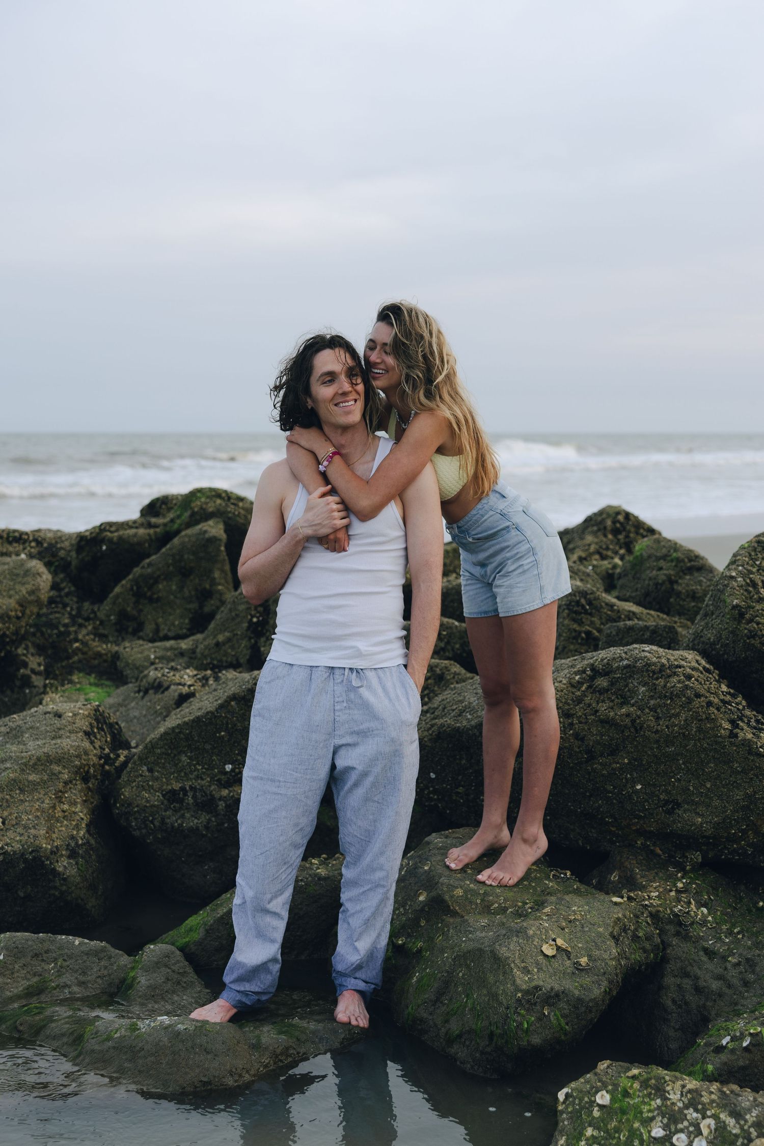 Two people share intimate moments while sitting on dark coastal rocks with ocean waves in the background.