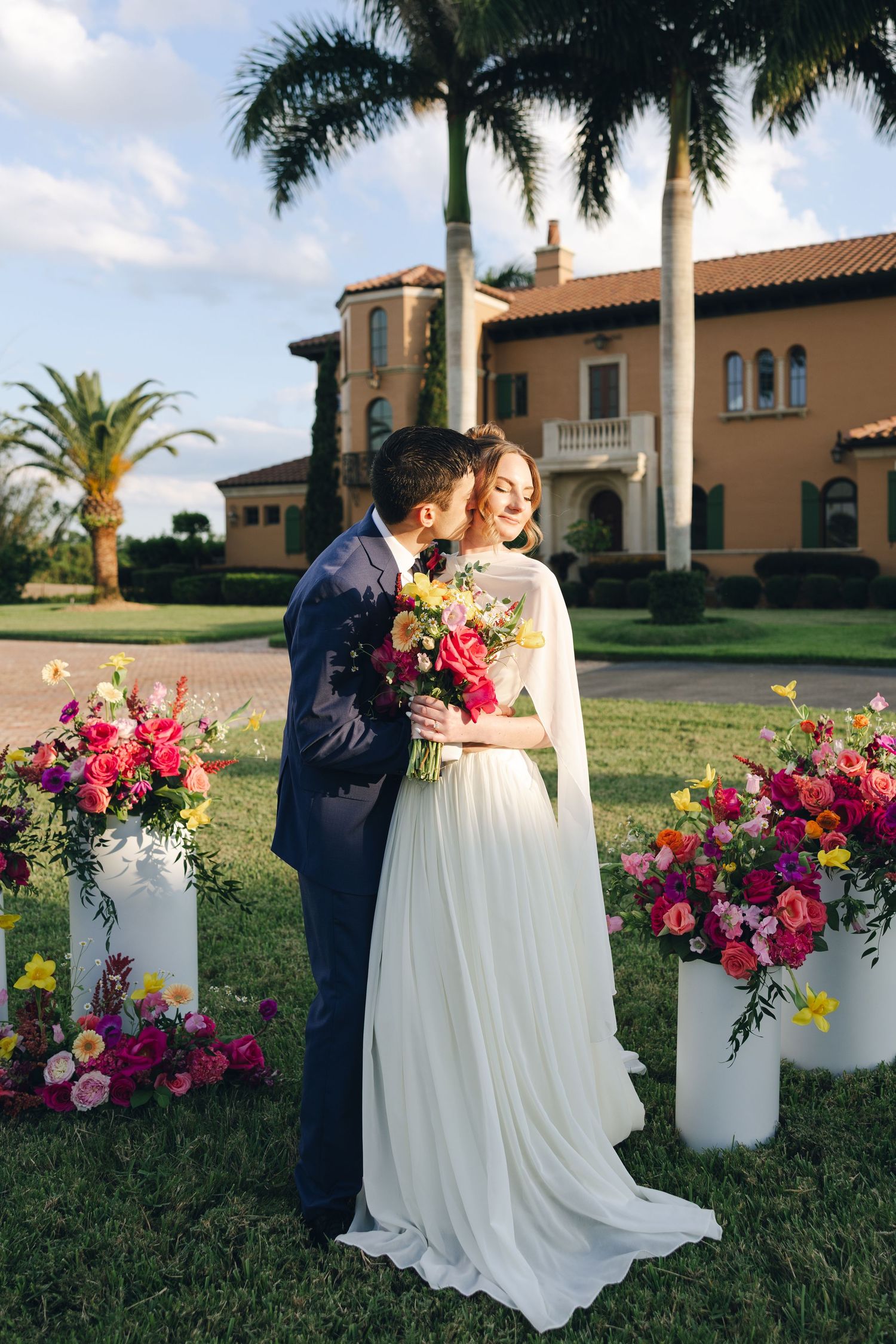 A couple in wedding attire embraces on a lawn with vibrant pink and yellow floral arrangements and palm trees.