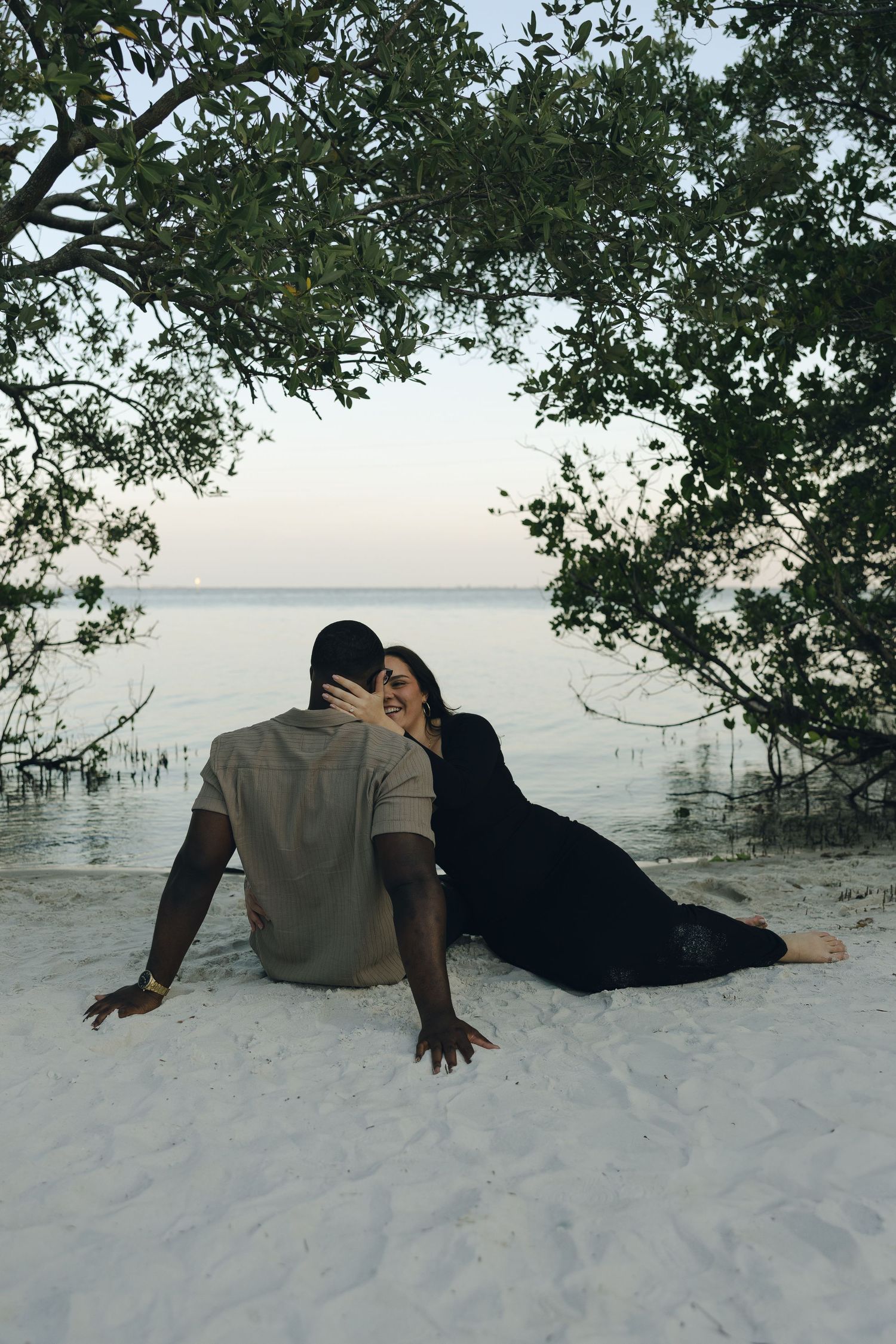 A romantic couple sits together on a white sandy beach at sunset with mangroves in the background.