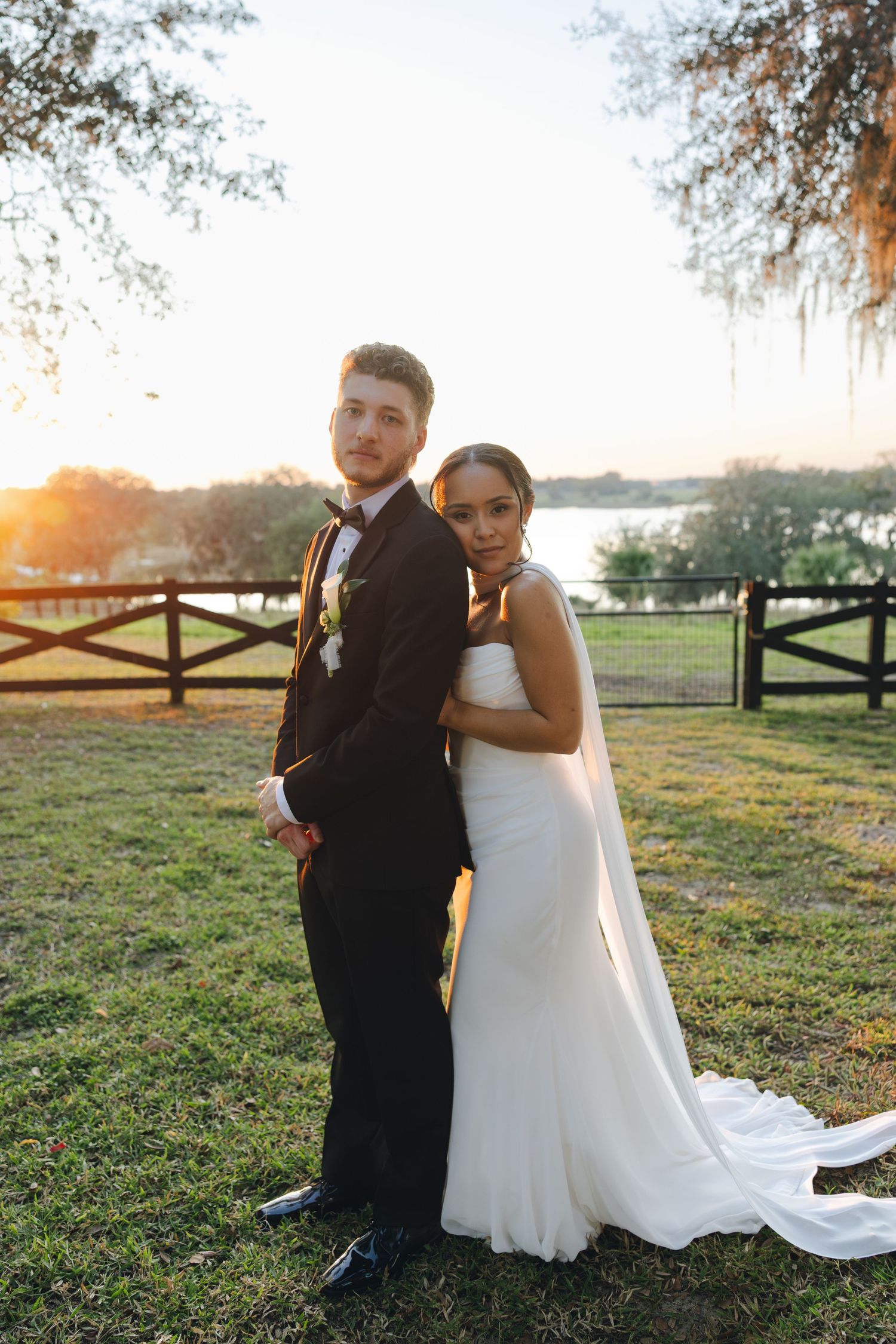 A couple in wedding attire sits together on grass at sunset with a rustic wooden fence in the background.
