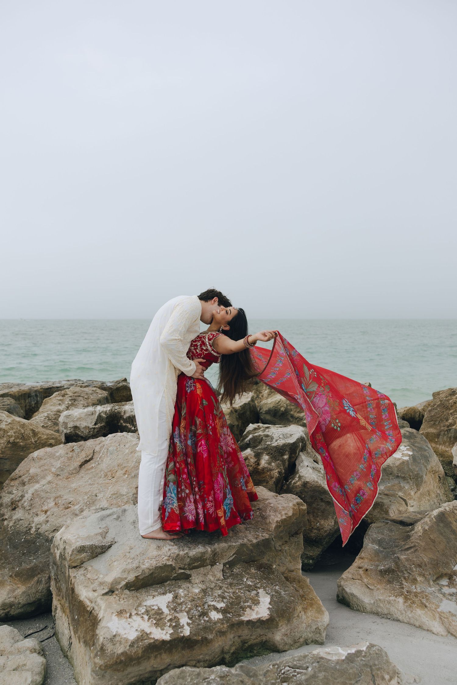A figure in white and red flowing fabric poses gracefully on coastal rocks against a misty ocean backdrop.