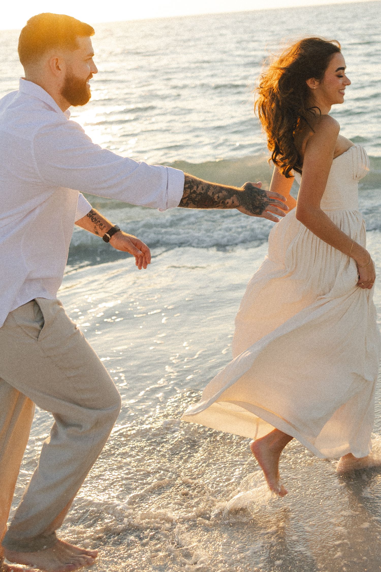 A couple runs playfully along the beach at sunset, with the woman in a flowing white dress and waves in the background.