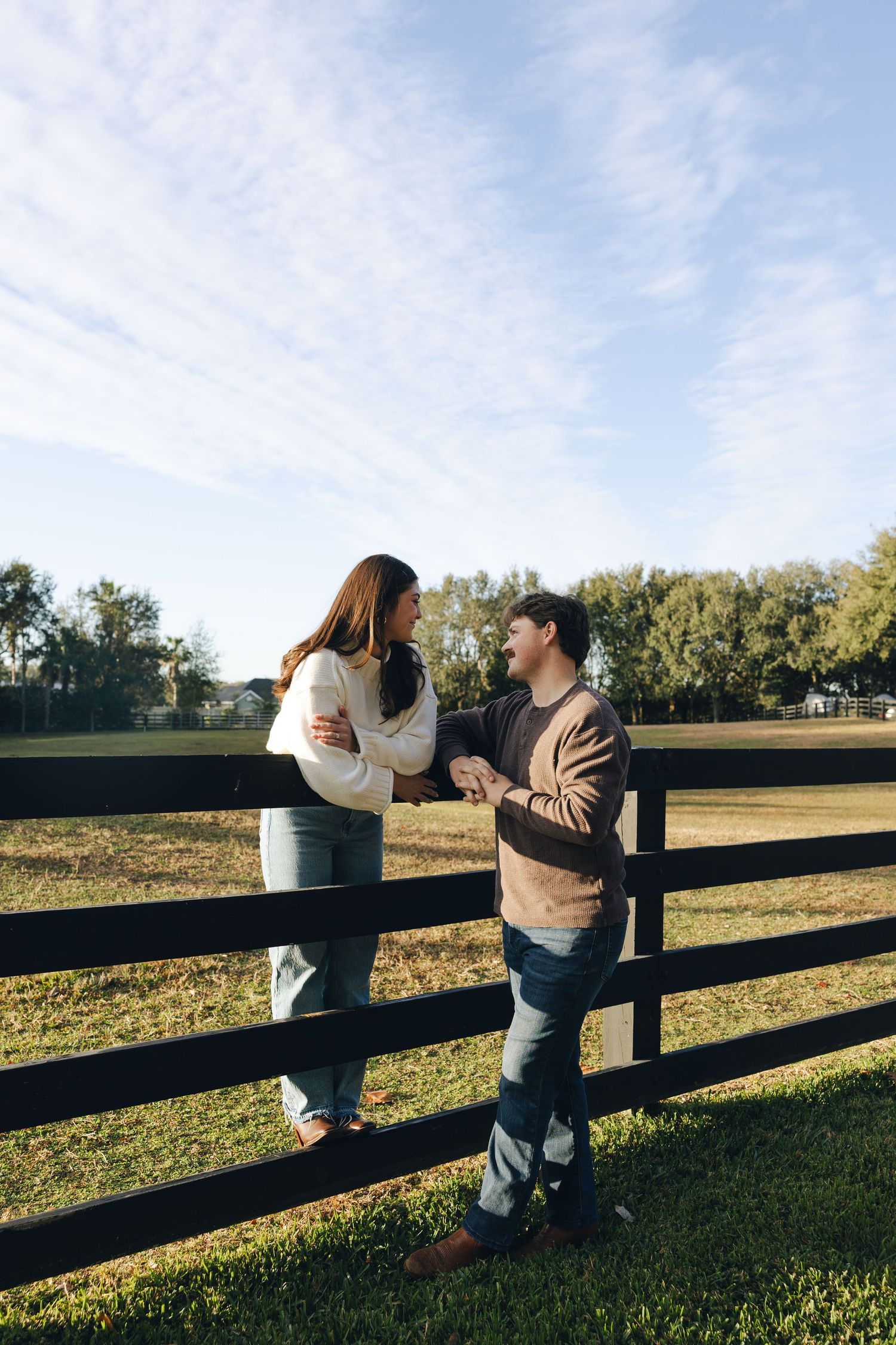 A couple leans on a wooden fence in a sunlit field as they share an intimate moment at sunset.
