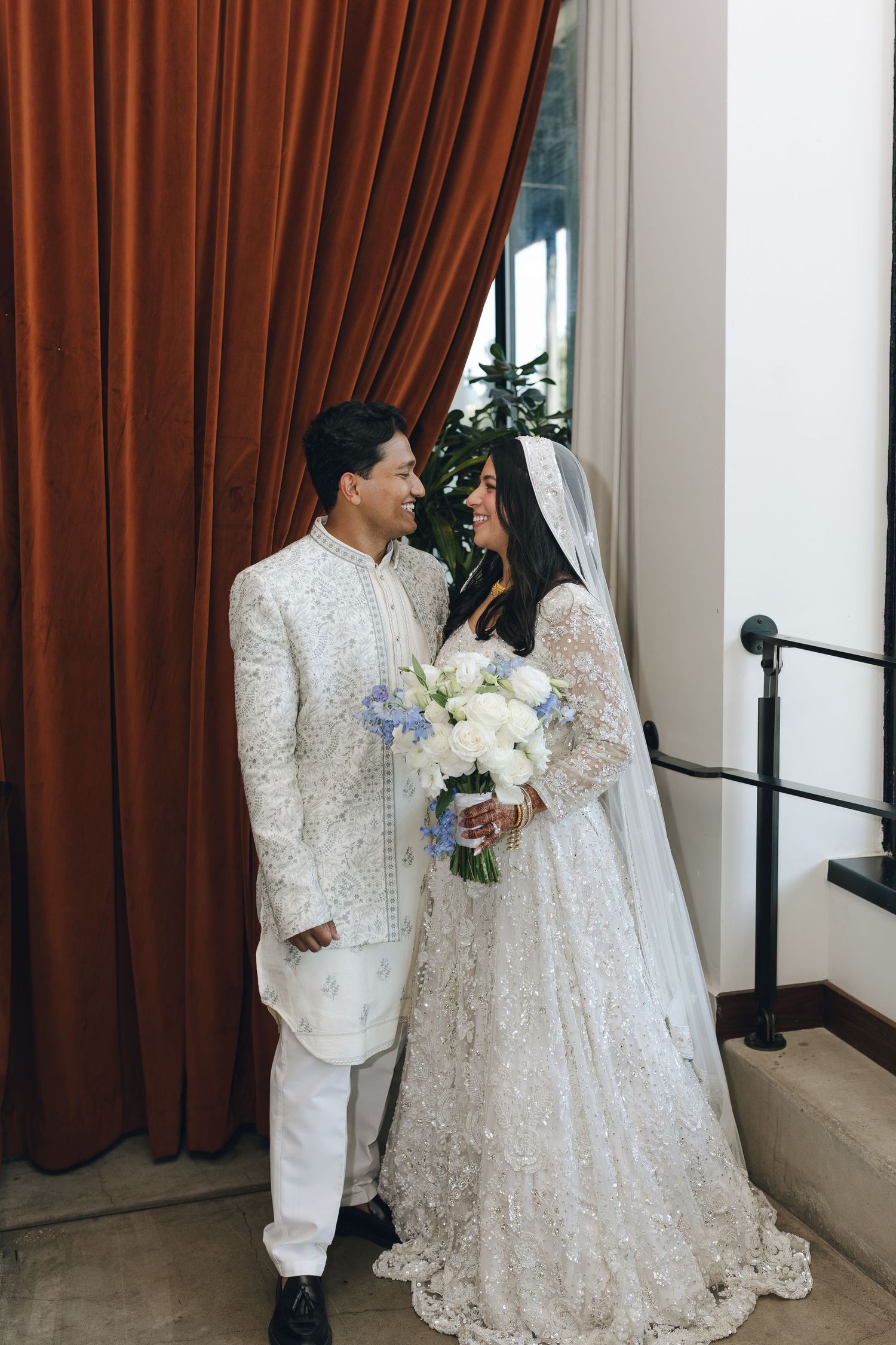 A couple in white wedding attire share an intimate moment against brown curtains and white walls during their ceremony.