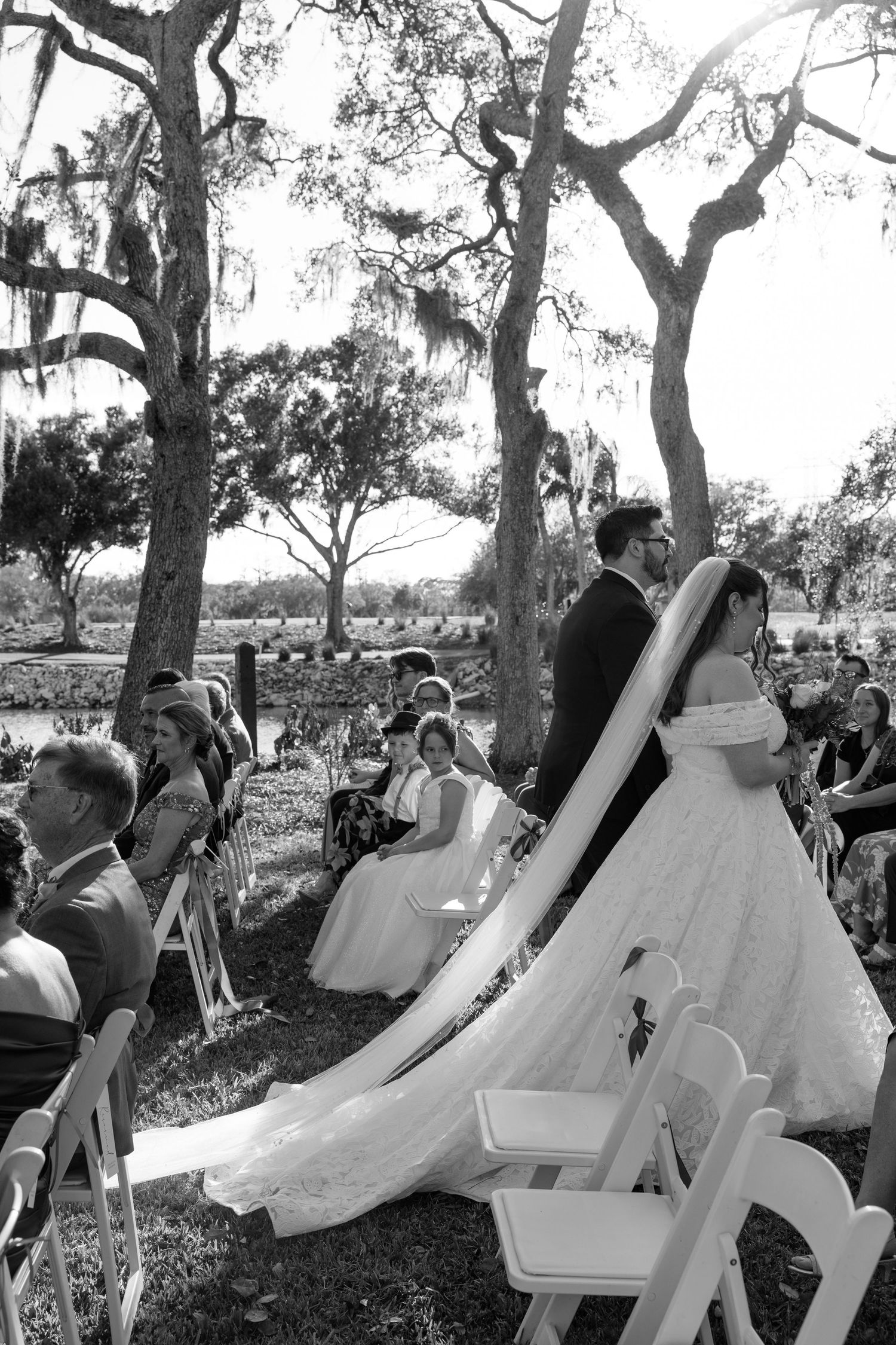 A vintage black and white photograph shows a group of people sitting outdoors under large oak trees.