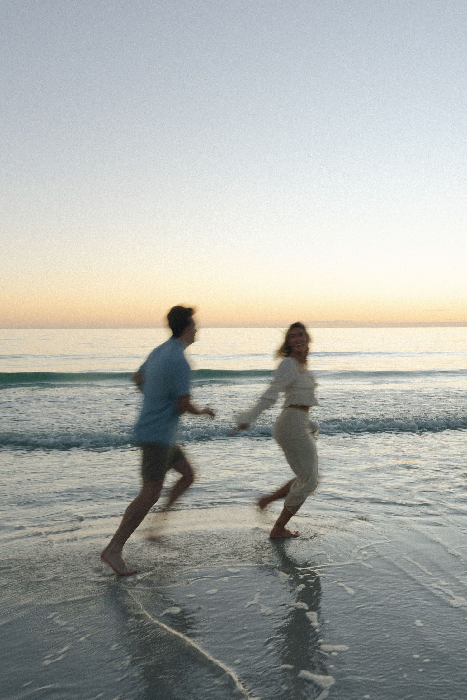 Couple playfully running and splashing through shallow ocean waves at sunset along a peaceful beach.