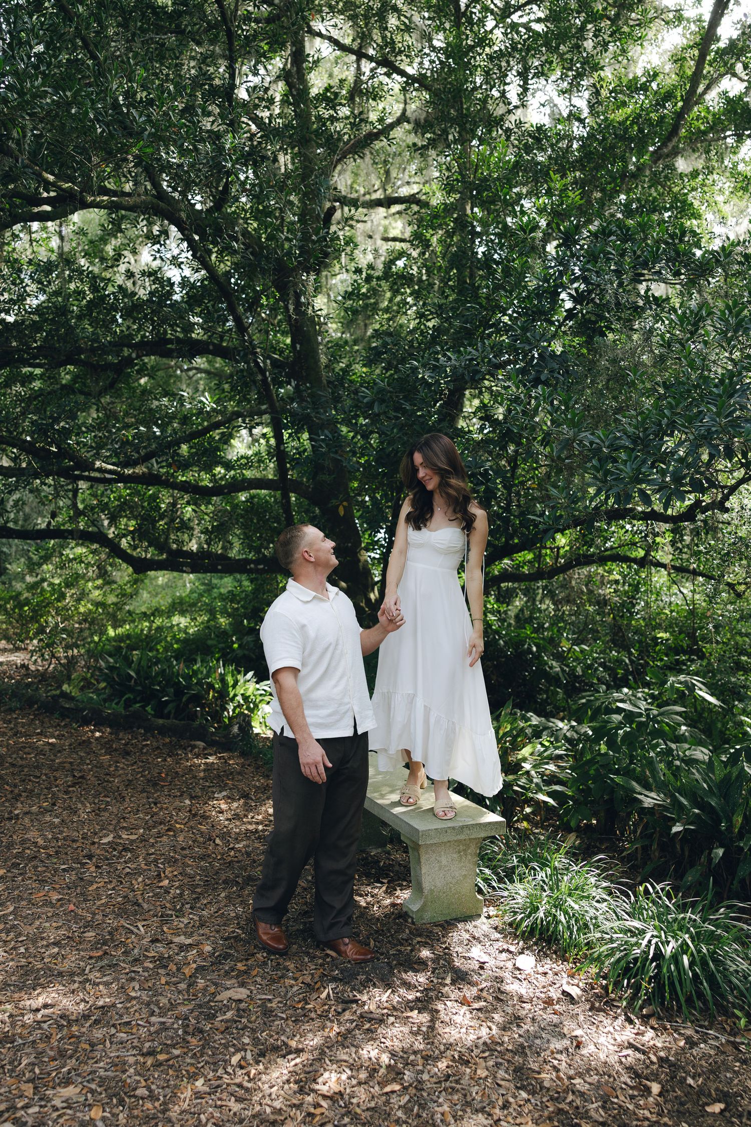 A couple in white and black attire poses together in a lush garden setting with dappled sunlight filtering through trees.