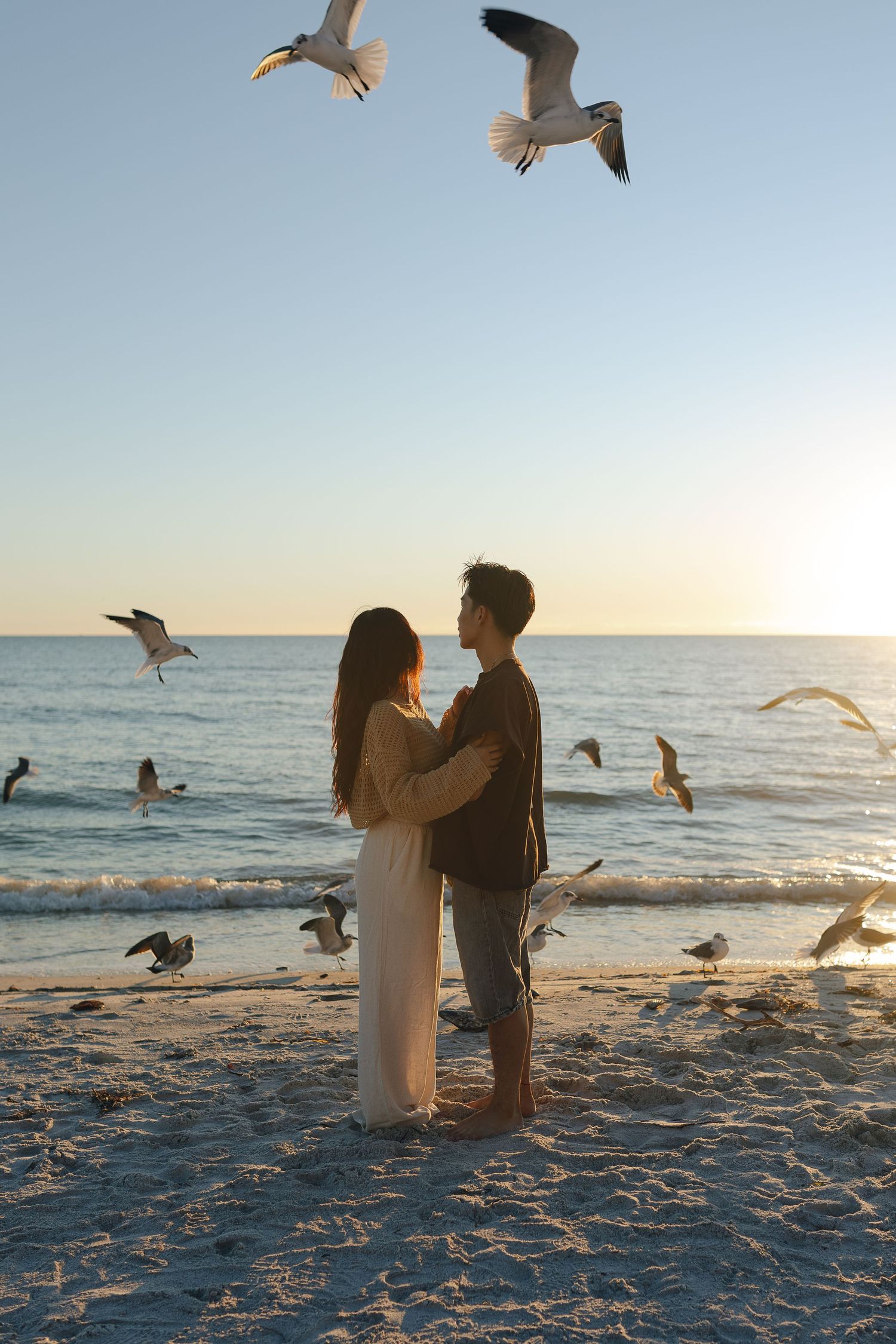 A couple stands on a beach at sunset surrounded by flying seagulls.