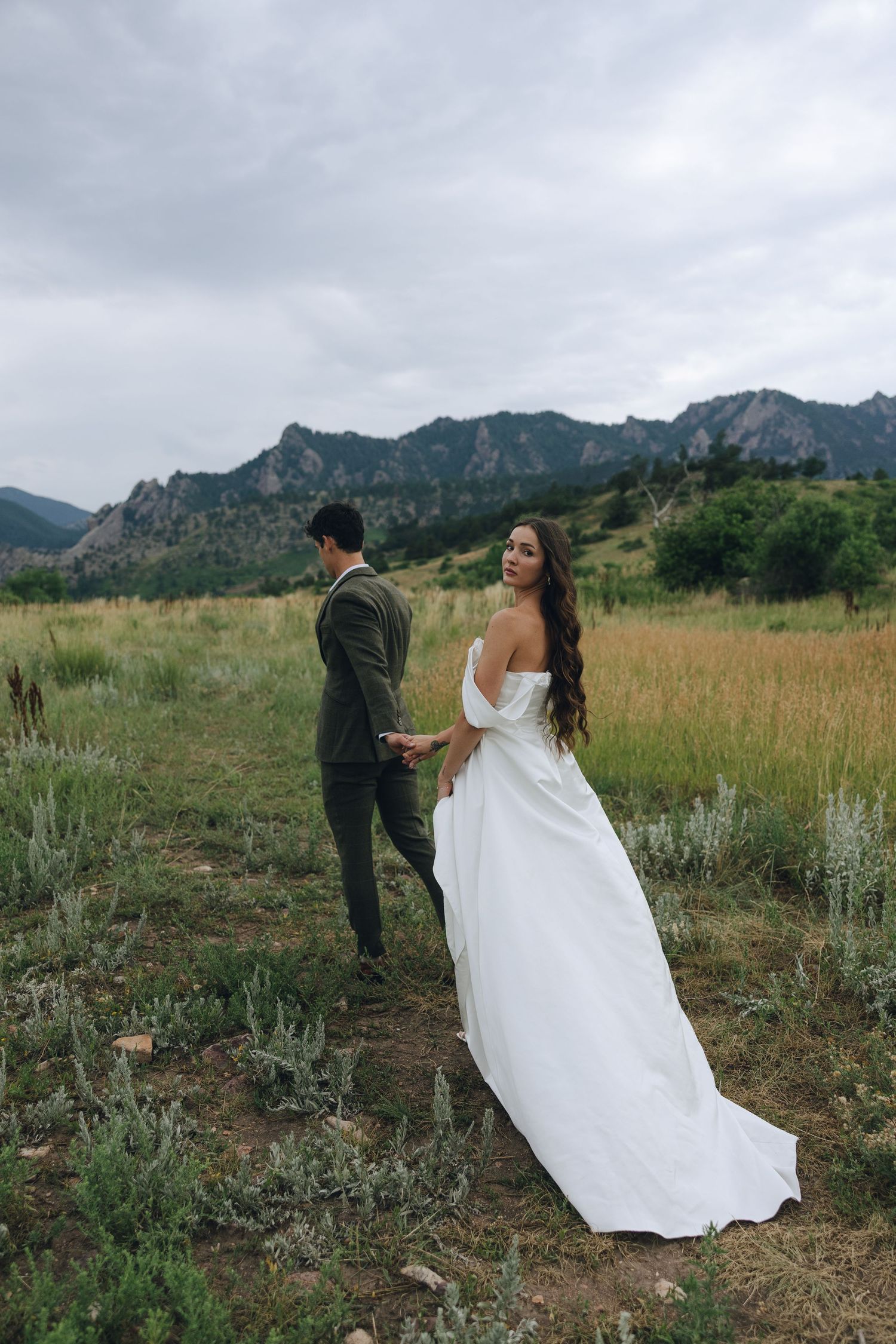 A couple in wedding attire stands in a meadow with dramatic mountain peaks and cloudy skies in the background.
