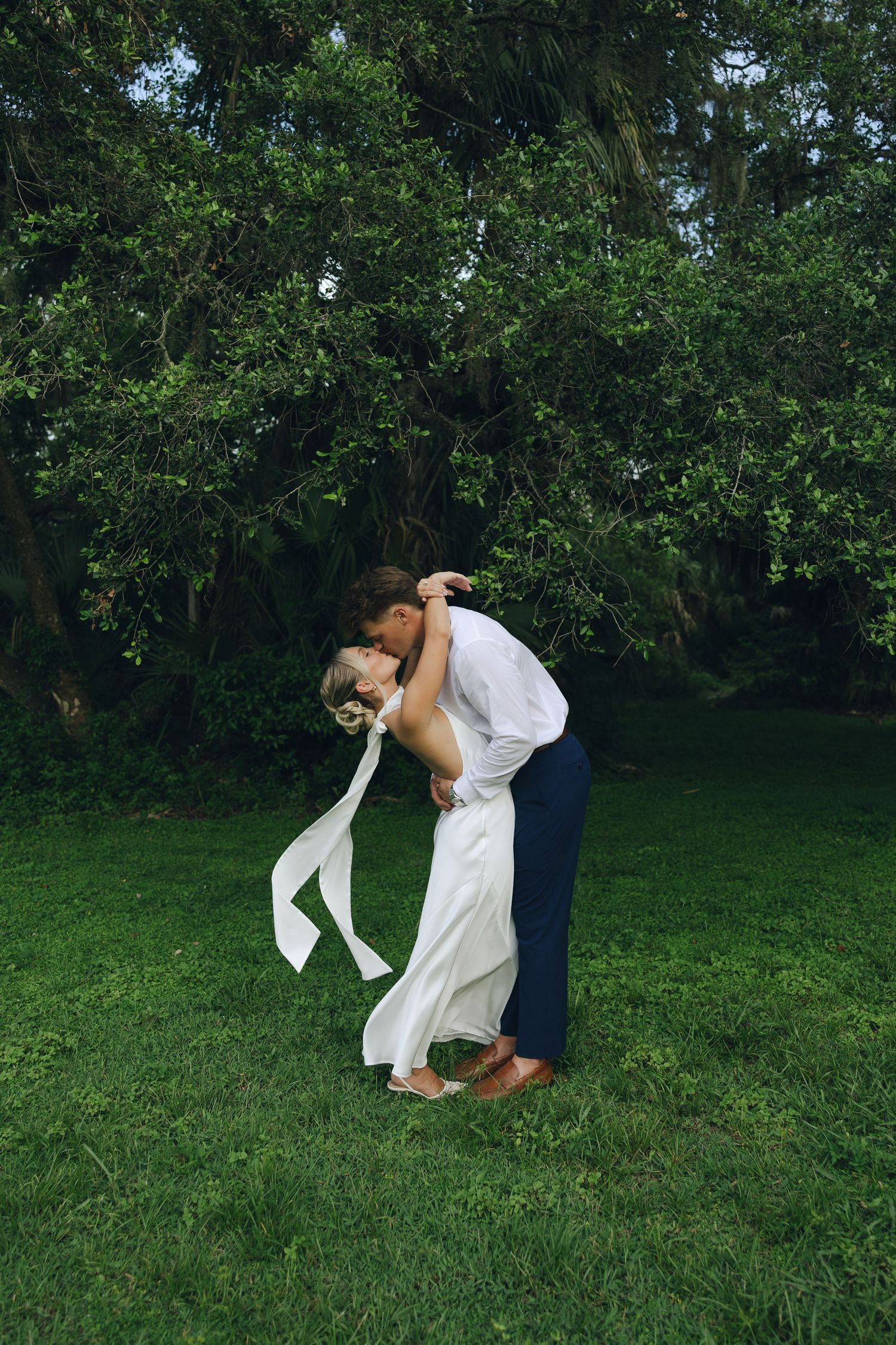 A couple shares a romantic dip kiss on a lush green lawn with trees in the background.