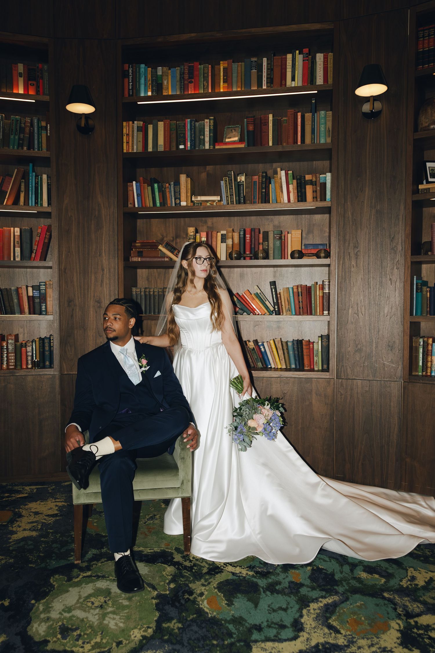 A bride and groom pose playfully in front of a wood-paneled library wall filled with books.