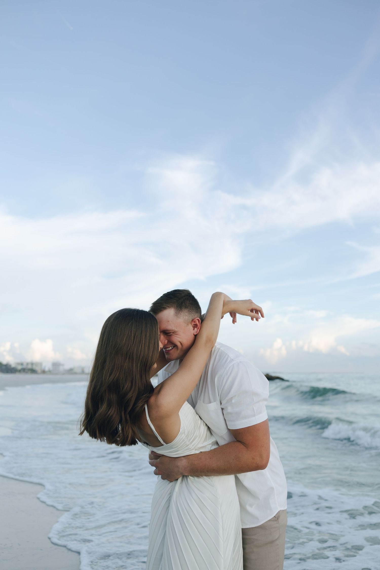 A romantic couple embraces on a beach at sunset with ocean waves and cloudy sky in the background.