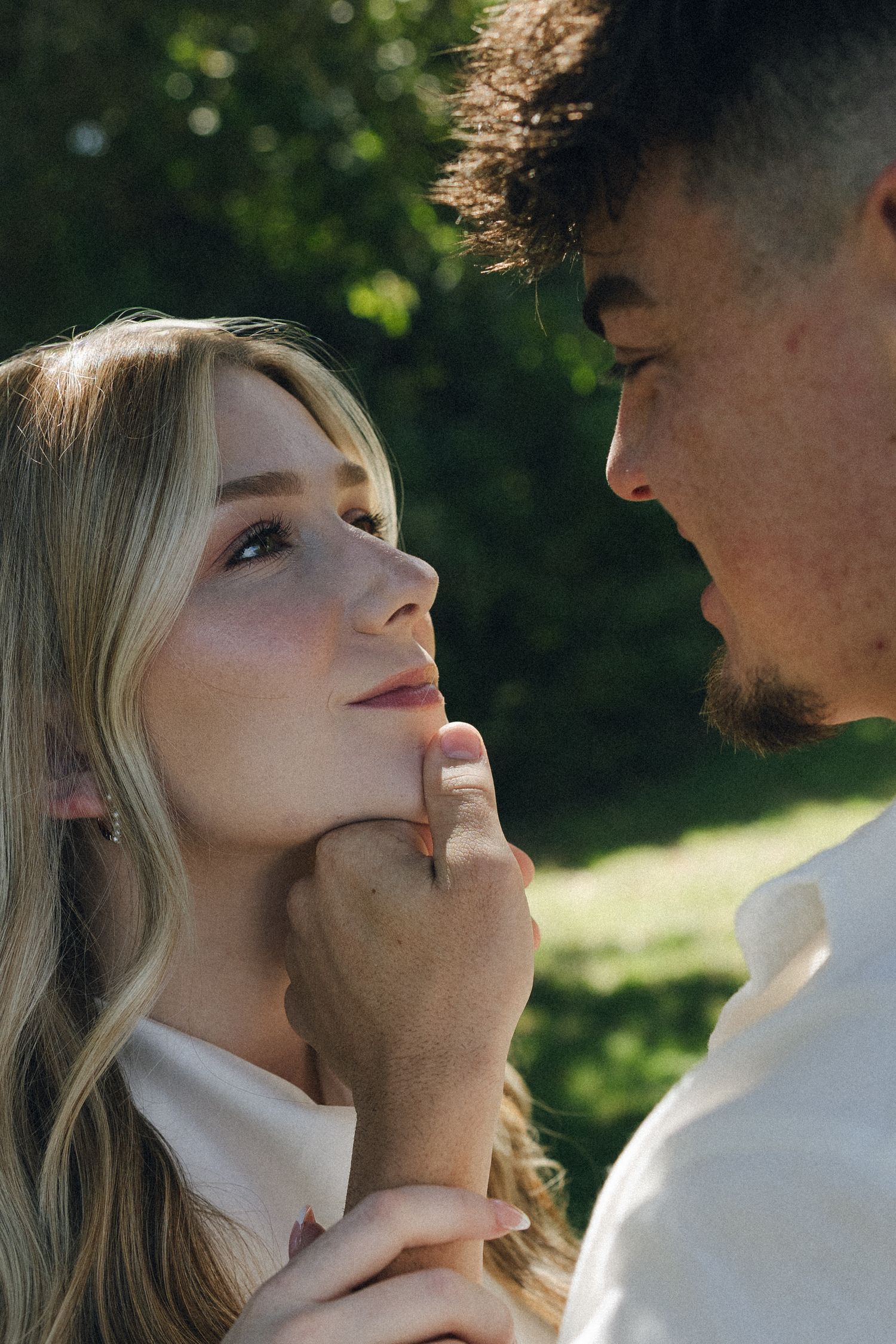 A romantic couple shares intimate moments in a sun-dappled outdoor setting wearing white clothing.