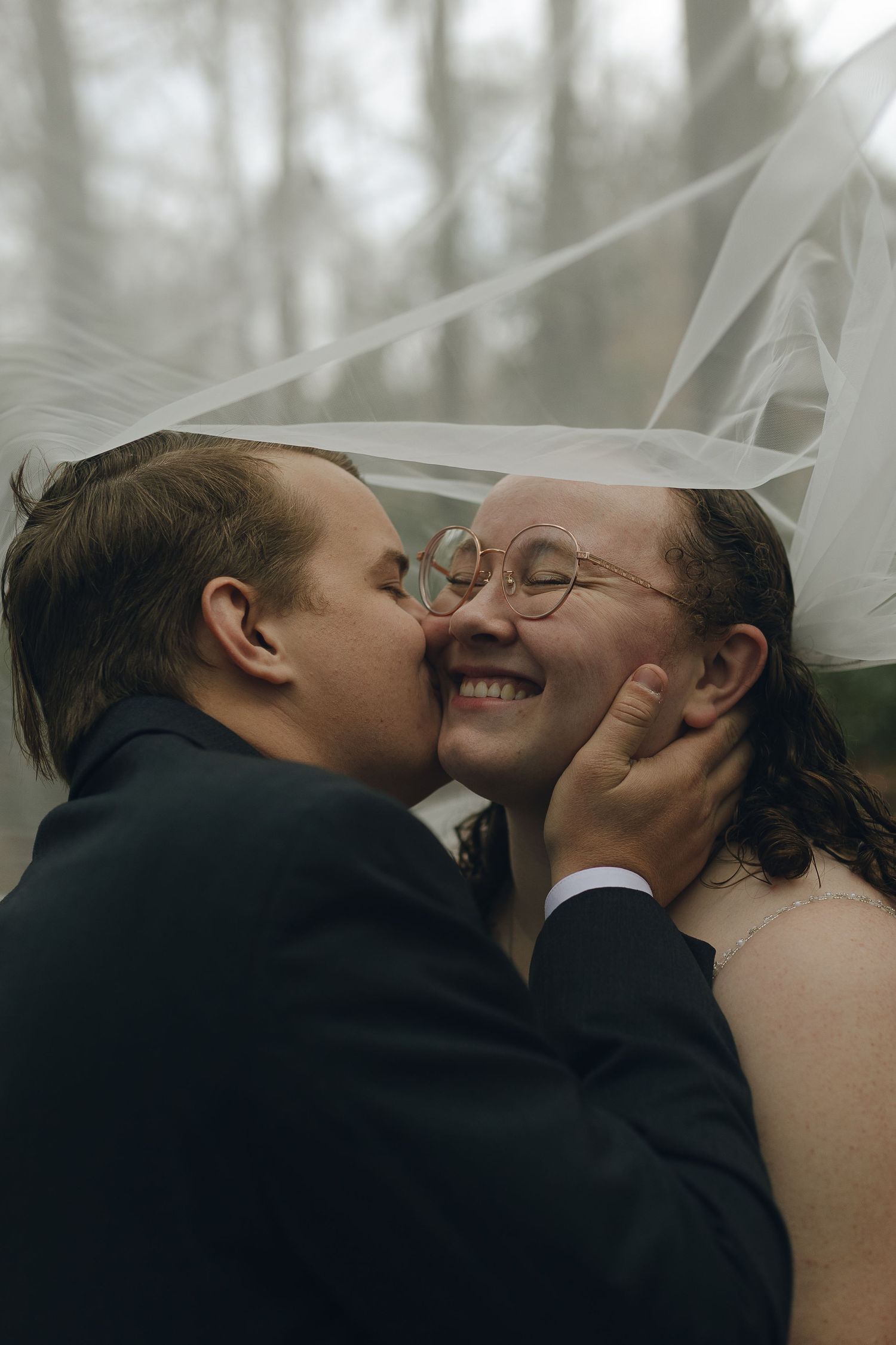 Intimate wedding moment captured under flowing veil in moody forest setting.