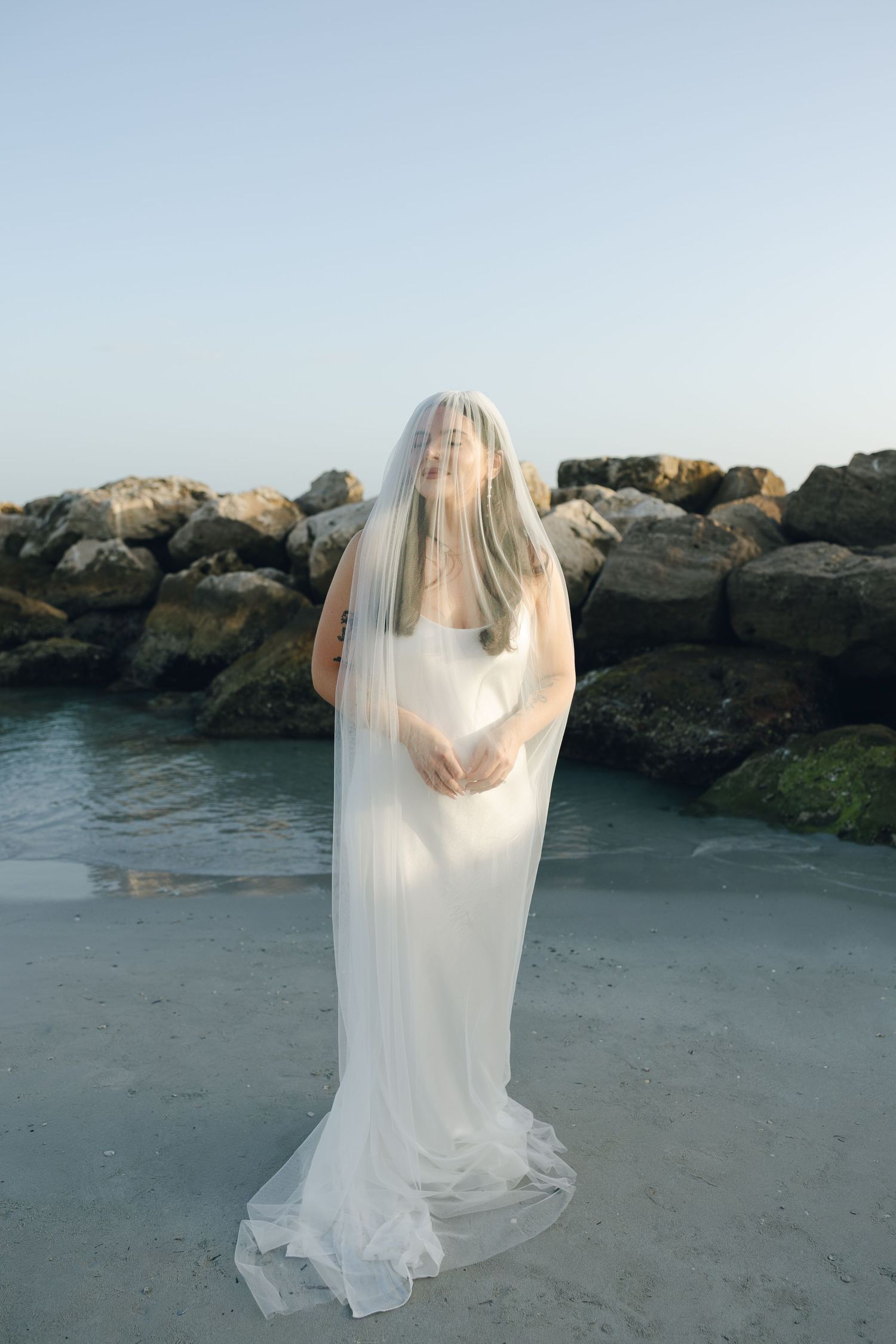 A figure in a flowing white wedding dress and veil stands on a misty beach with rocky outcroppings in the background.