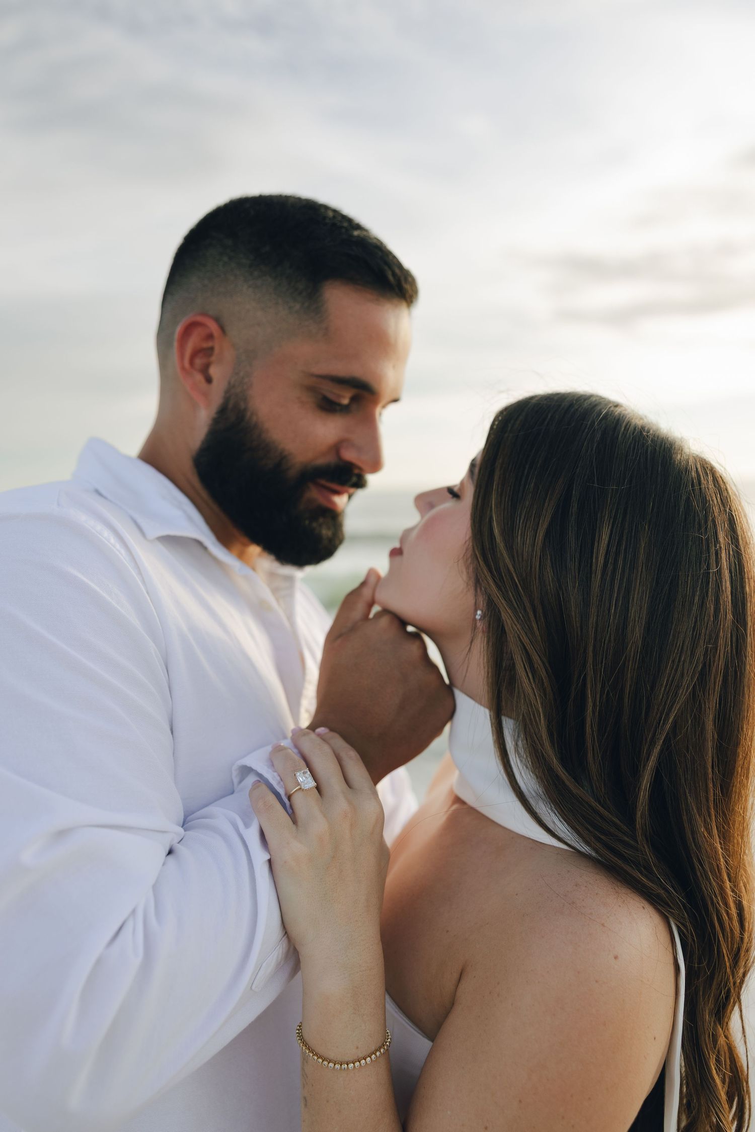 Romantic couple in white embraces and kisses against cloudy sky background in series of intimate photos.