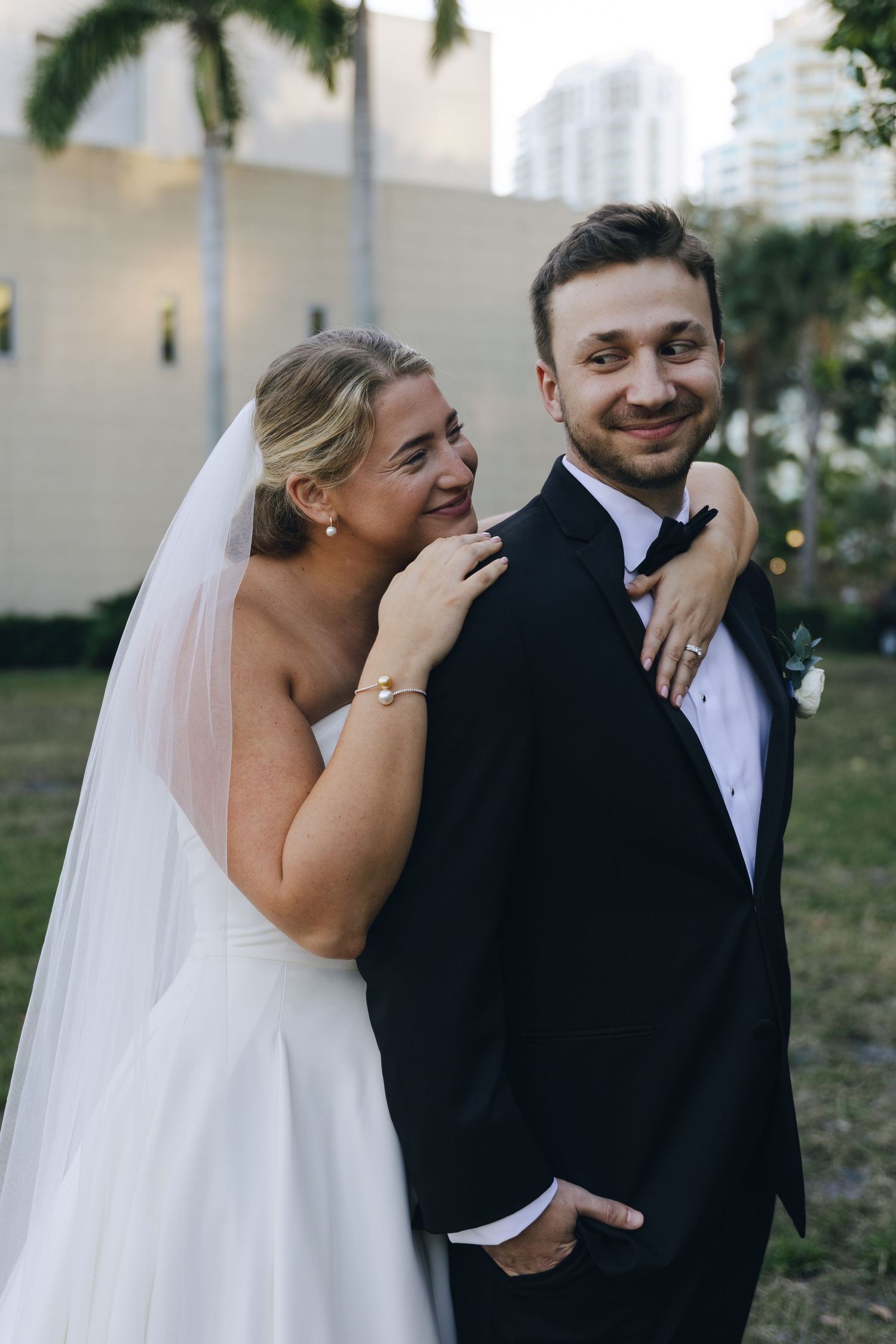 A joyful wedding moment captured outdoors with palm trees, showing a bride in white embracing her groom in a black tuxedo.