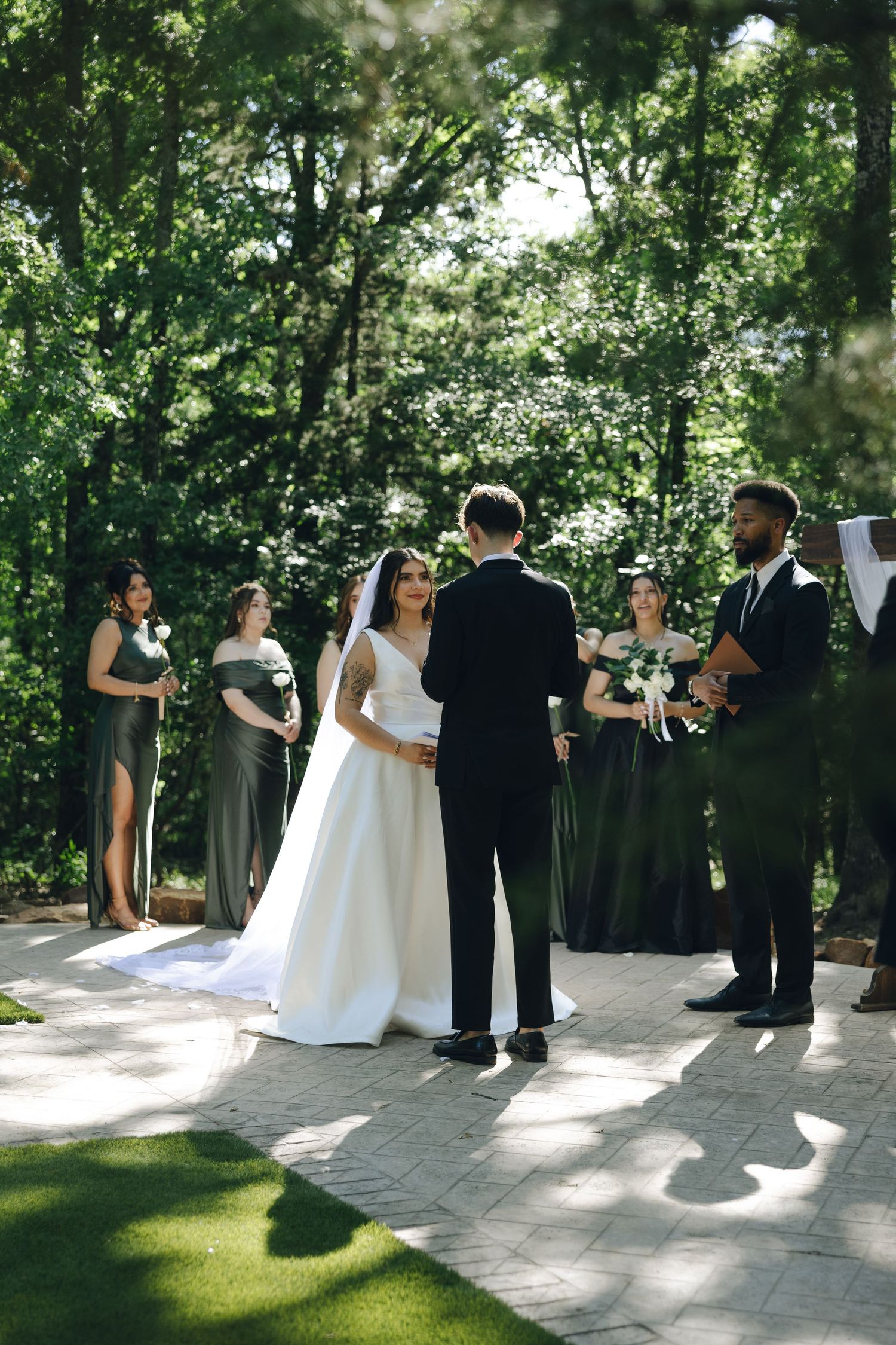 An outdoor wedding ceremony takes place in a serene garden setting with greenery and dappled sunlight on stone pavement.