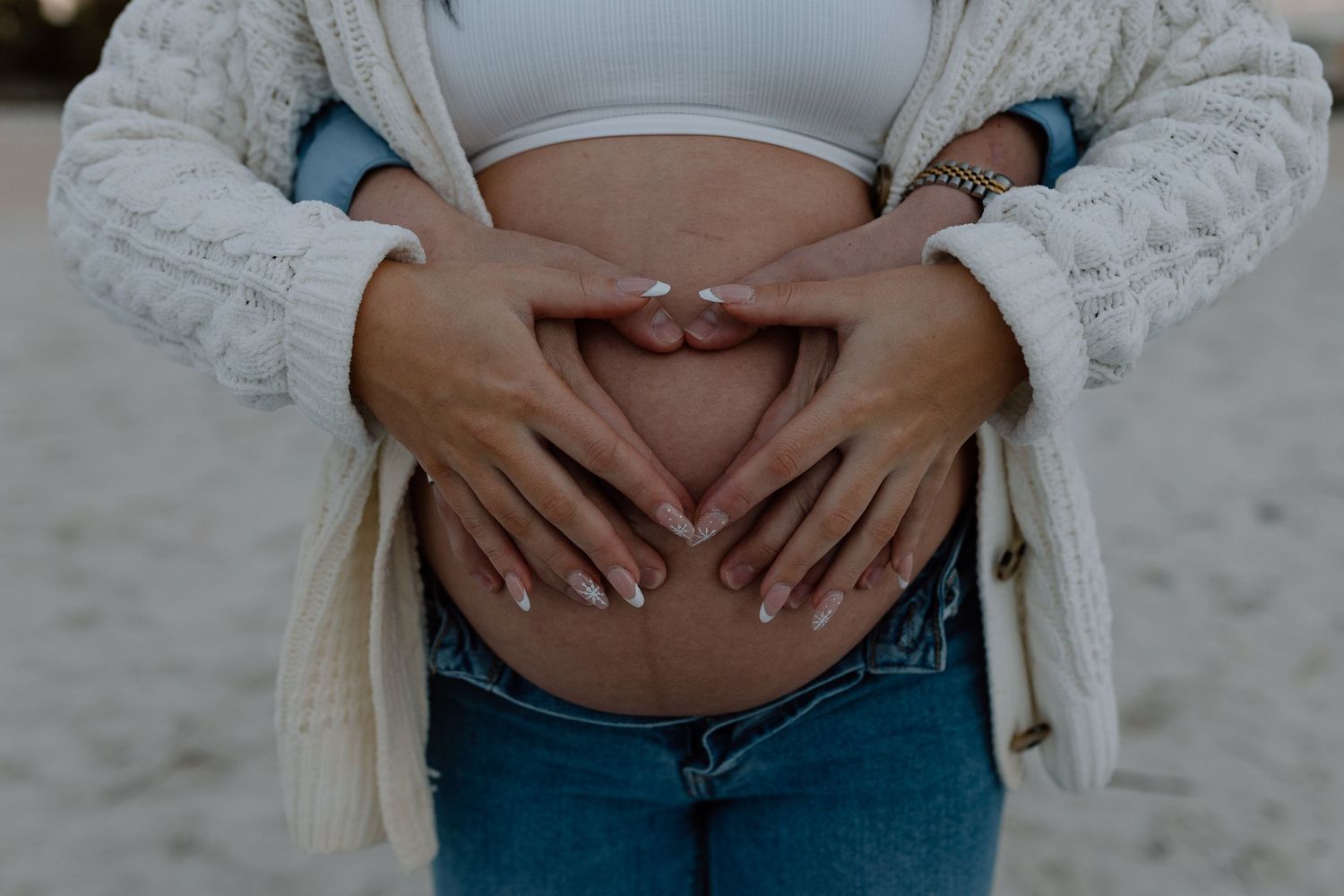 Hands form a heart shape over a pregnant belly wearing jeans and a white crop top.