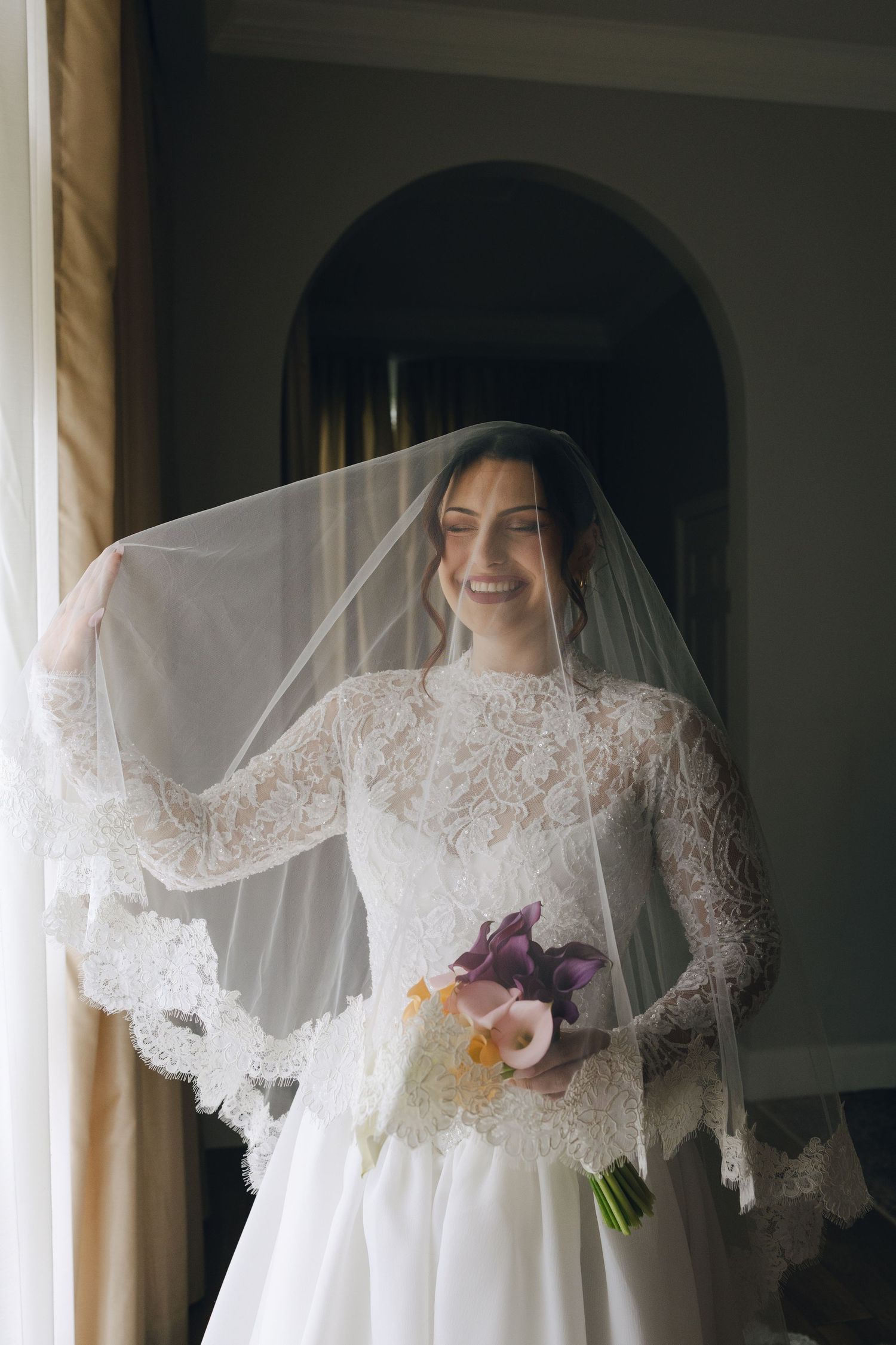 A bride in a vintage-style white lace wedding dress with long sleeves and veil stands in soft window light.