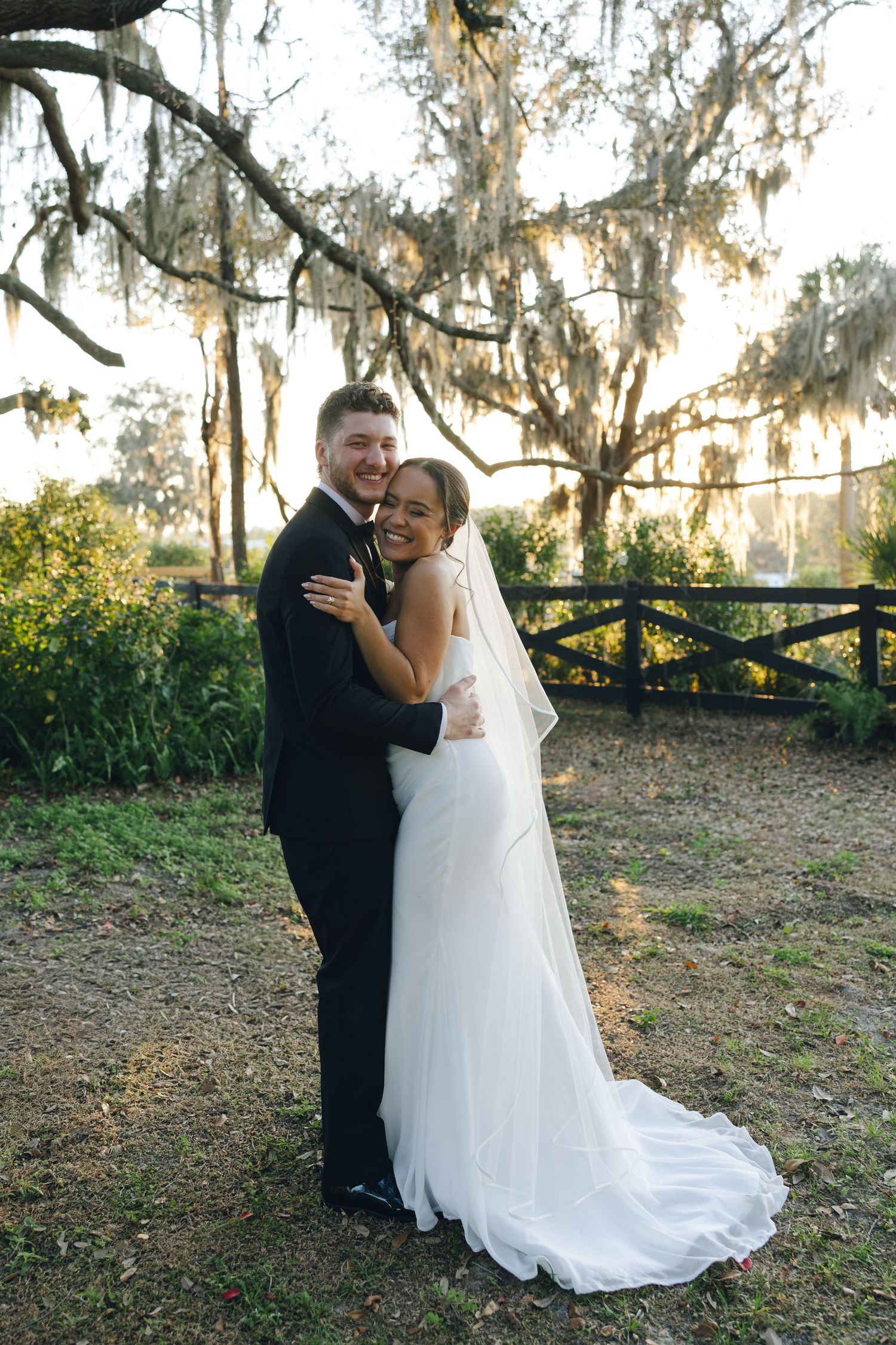 A couple shares a romantic moment under moss-draped trees at sunset during their wedding celebration.