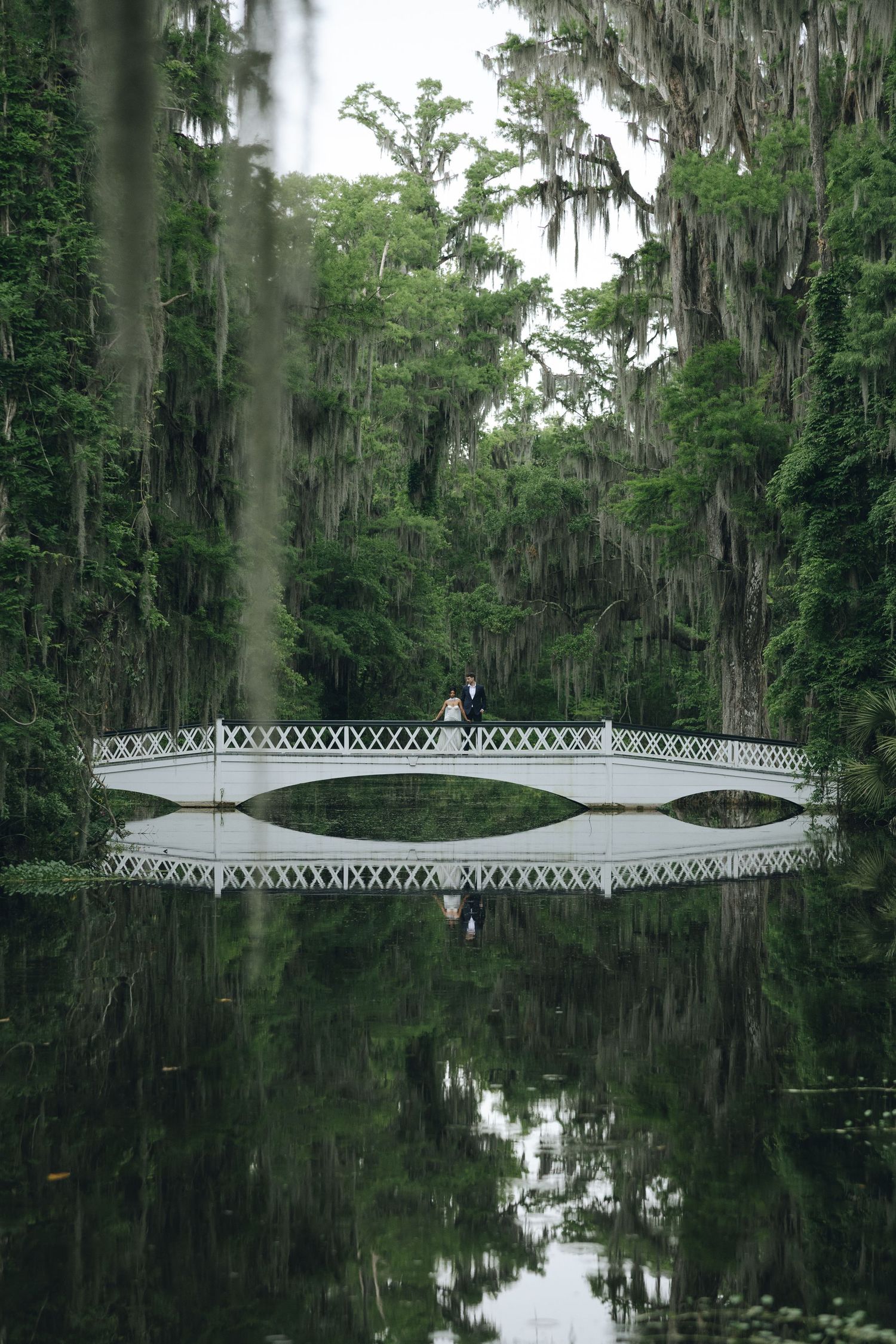 White ornate bridge reflects perfectly in still waters surrounded by Spanish moss covered trees.