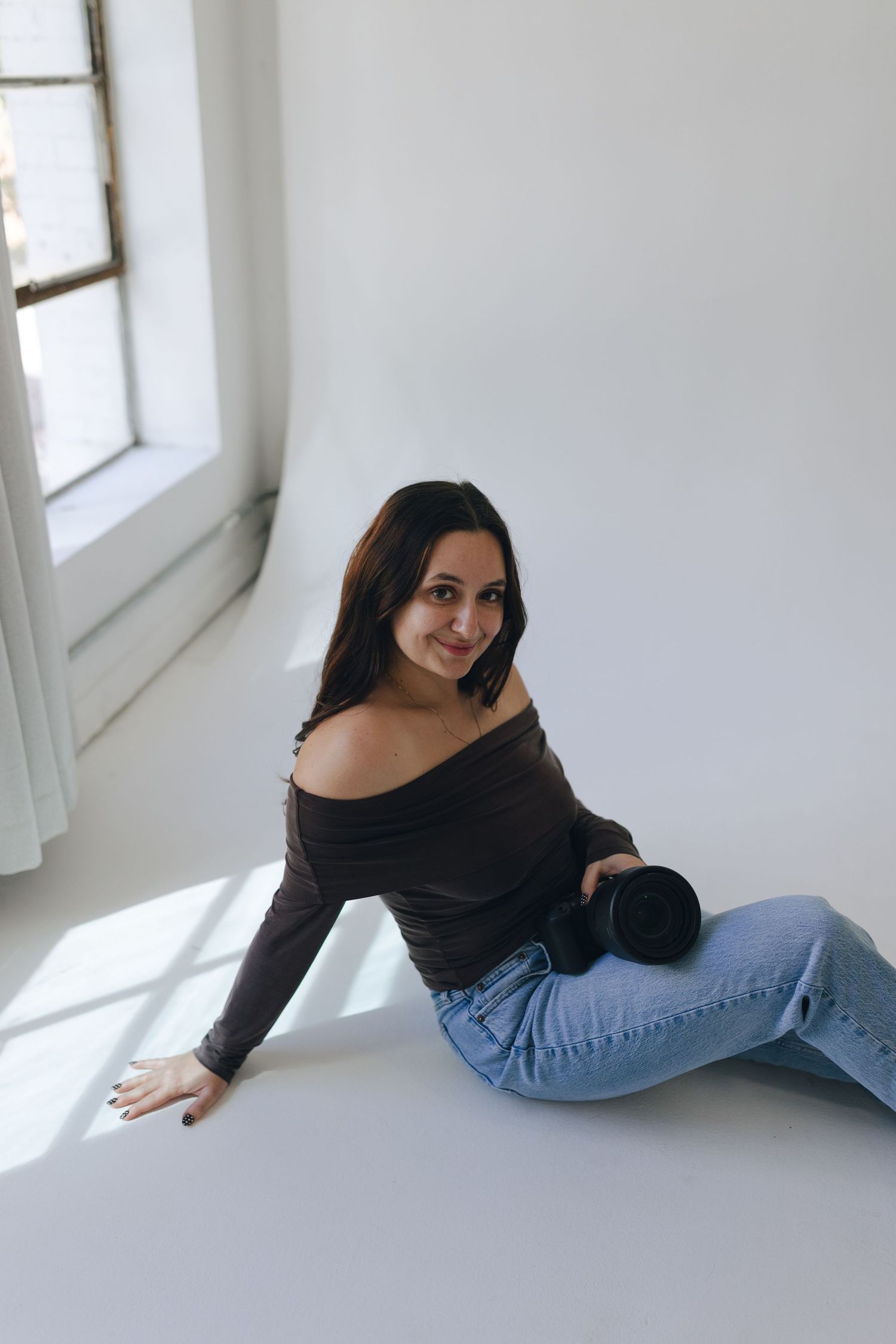 Woman in black off-shoulder top and blue jeans sits casually on white floor near window with natural lighting.