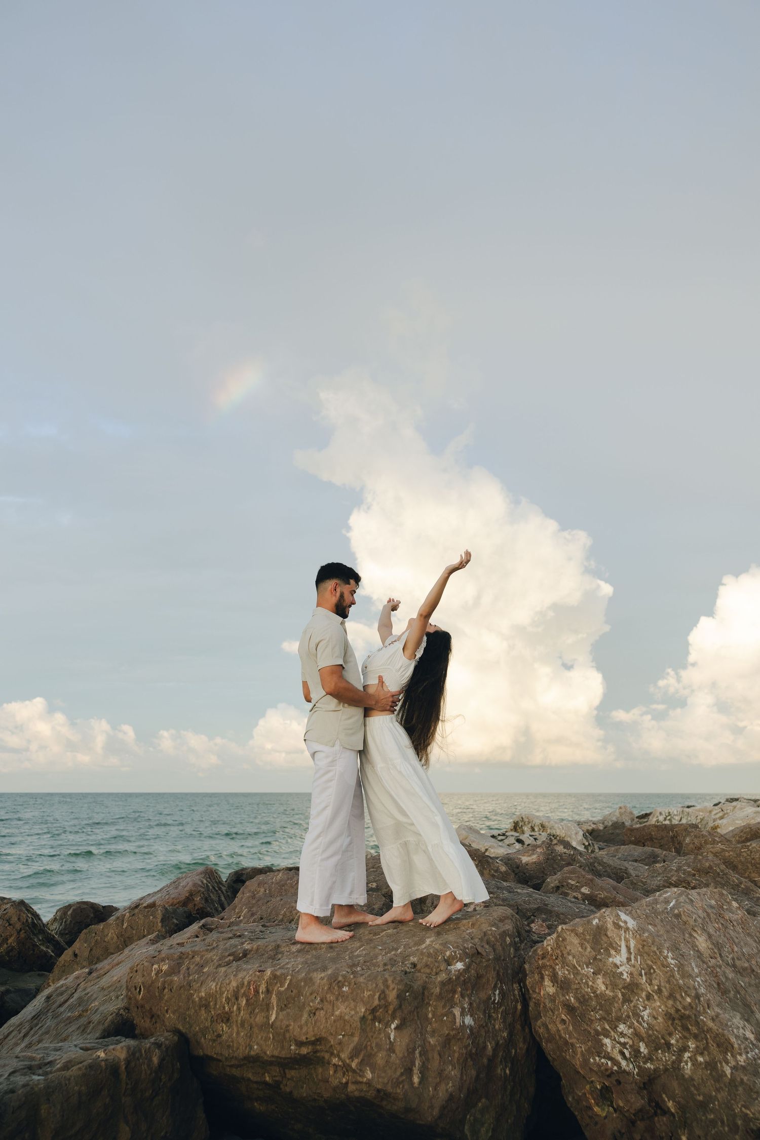 A couple in white attire poses romantically on coastal rocks with dramatic cloudy skies and ocean in the background.