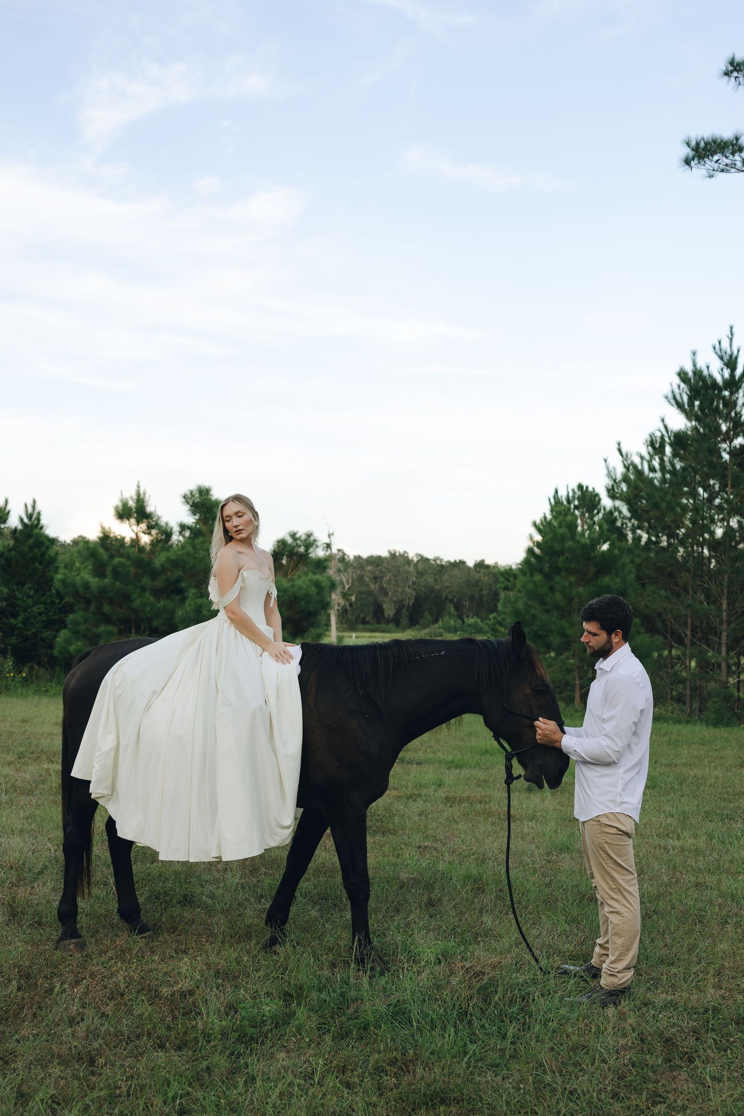 A couple poses playfully with a black horse in a grassy field during an outdoor photo session.