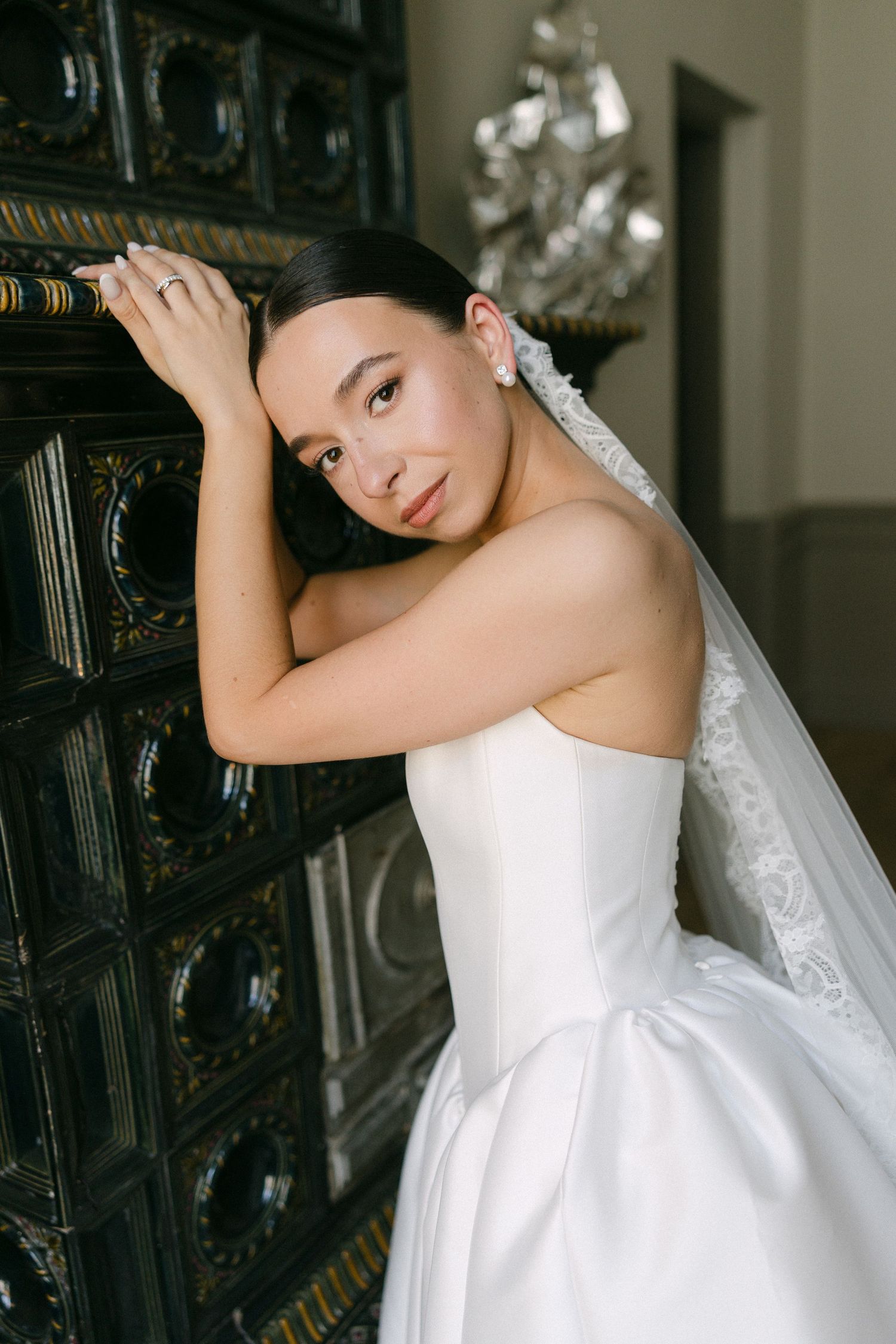 Elegant bride in a strapless white ballgown leaning against an ornate dark cabinet with a lace veil.