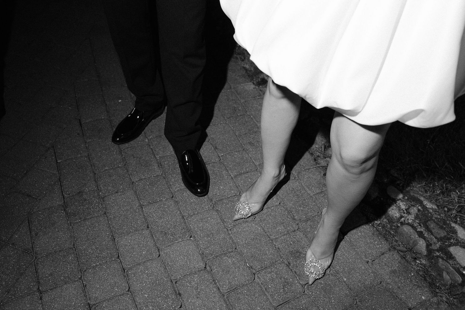 Black and white photo of a bride and groom's feet, showcasing elegant heels and dress shoes on cobblestone.