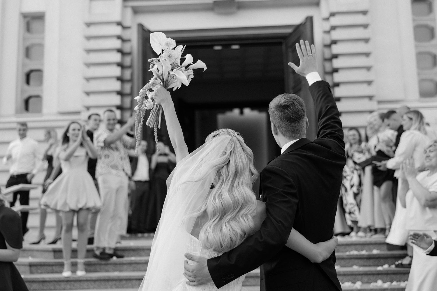 Black and white photo of newlyweds embracing on church steps as guests cheer and bride raises her white bouquet.