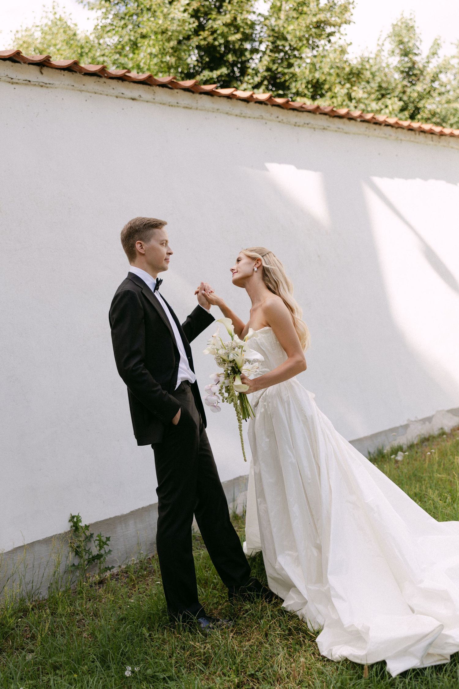 Bride and groom holding hands near a white wall in a sunlit garden, bride holding white bouquet.