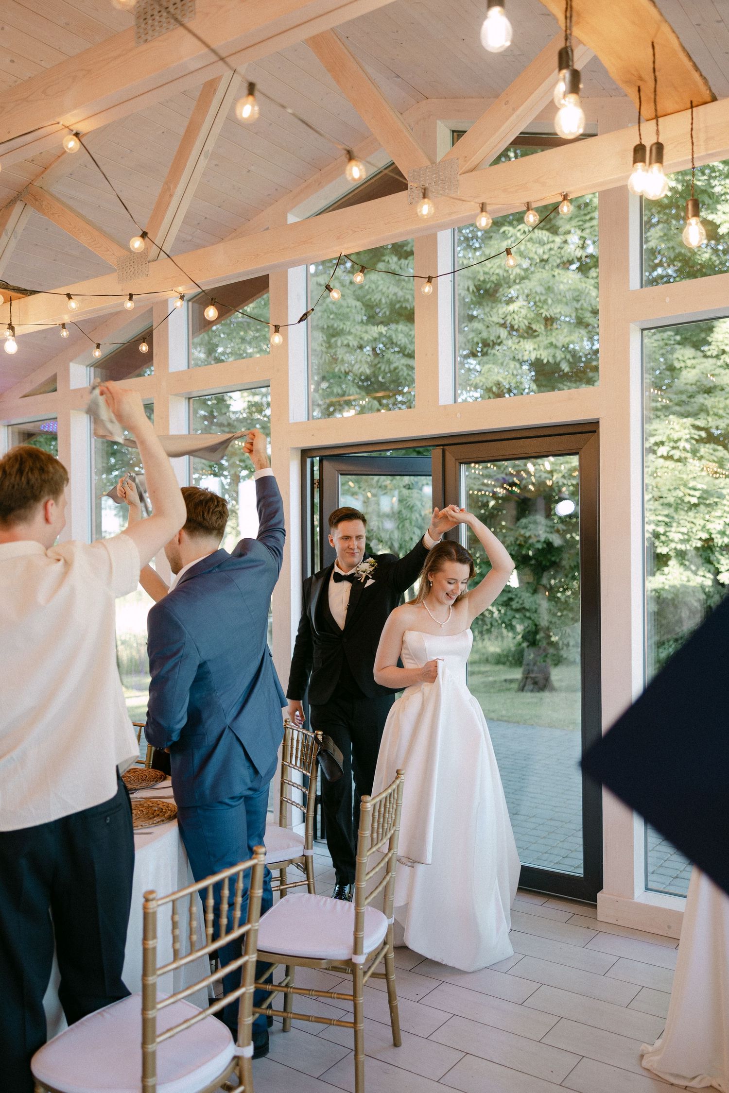 Bride and groom make grand entrance into elegant wedding reception venue with string lights and large windows.