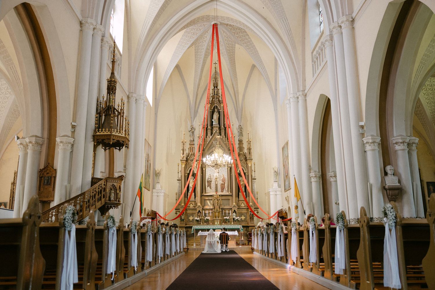 Elegant Catholic church interior with red ribbon decorations, white draping, and ornate golden altar.