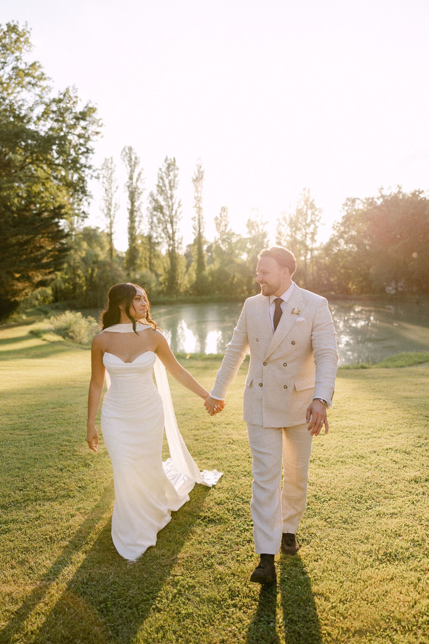 Bride and groom walking hand in hand at golden hour near a serene pond surrounded by trees.