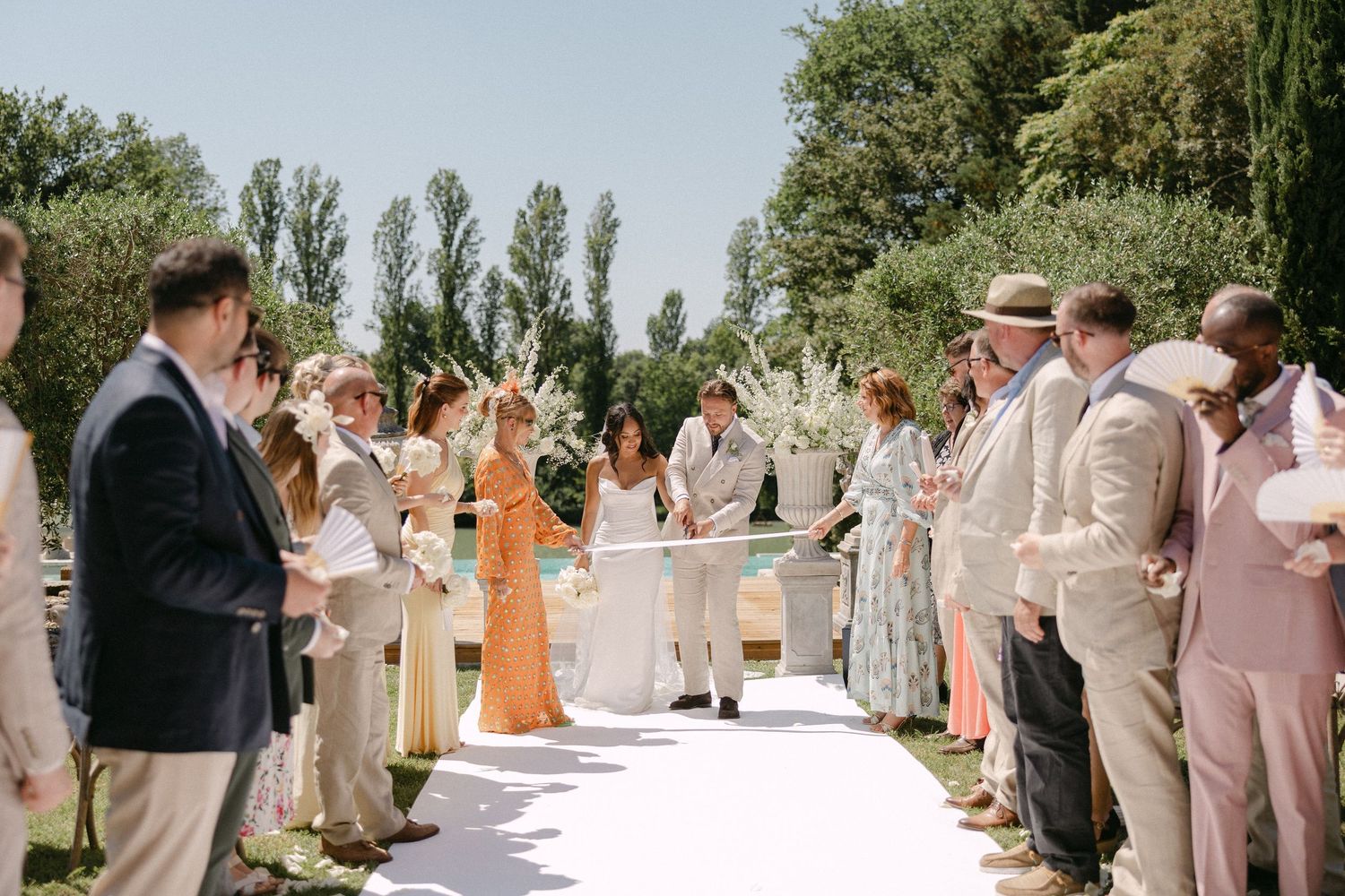 Wedding guests line white aisle as bride and groom walk through outdoor garden ceremony on sunny day.