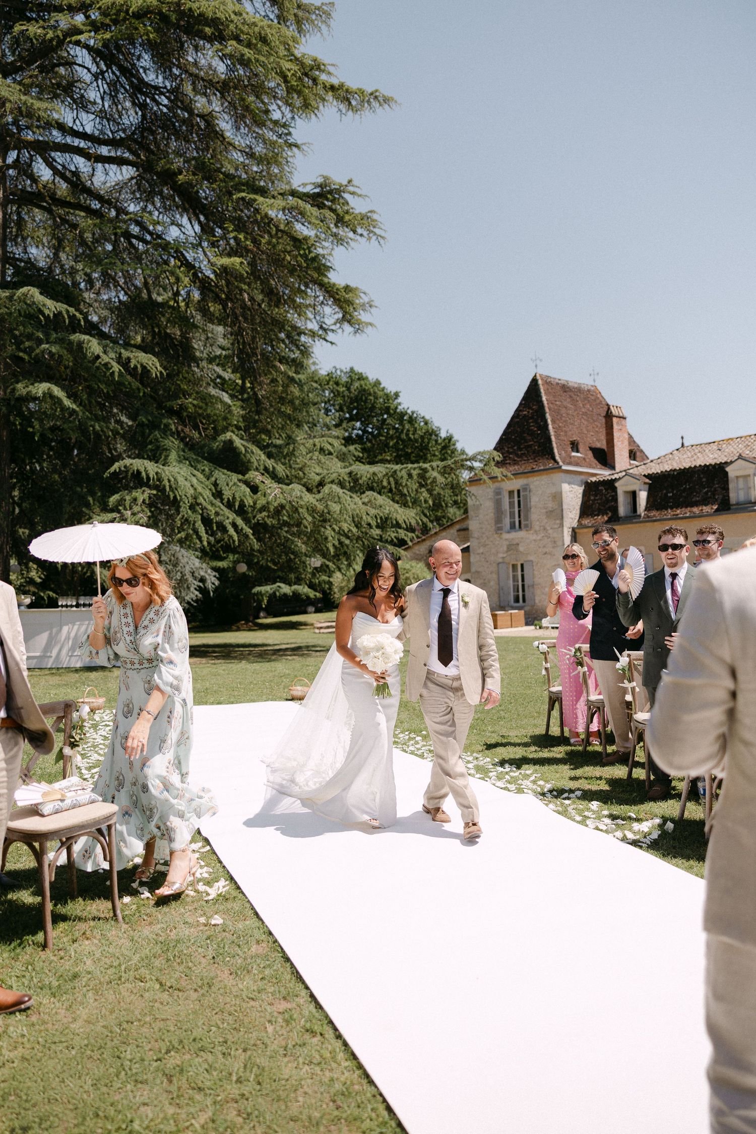 Bride walking down outdoor aisle with escort at sunny French chateau garden wedding ceremony.