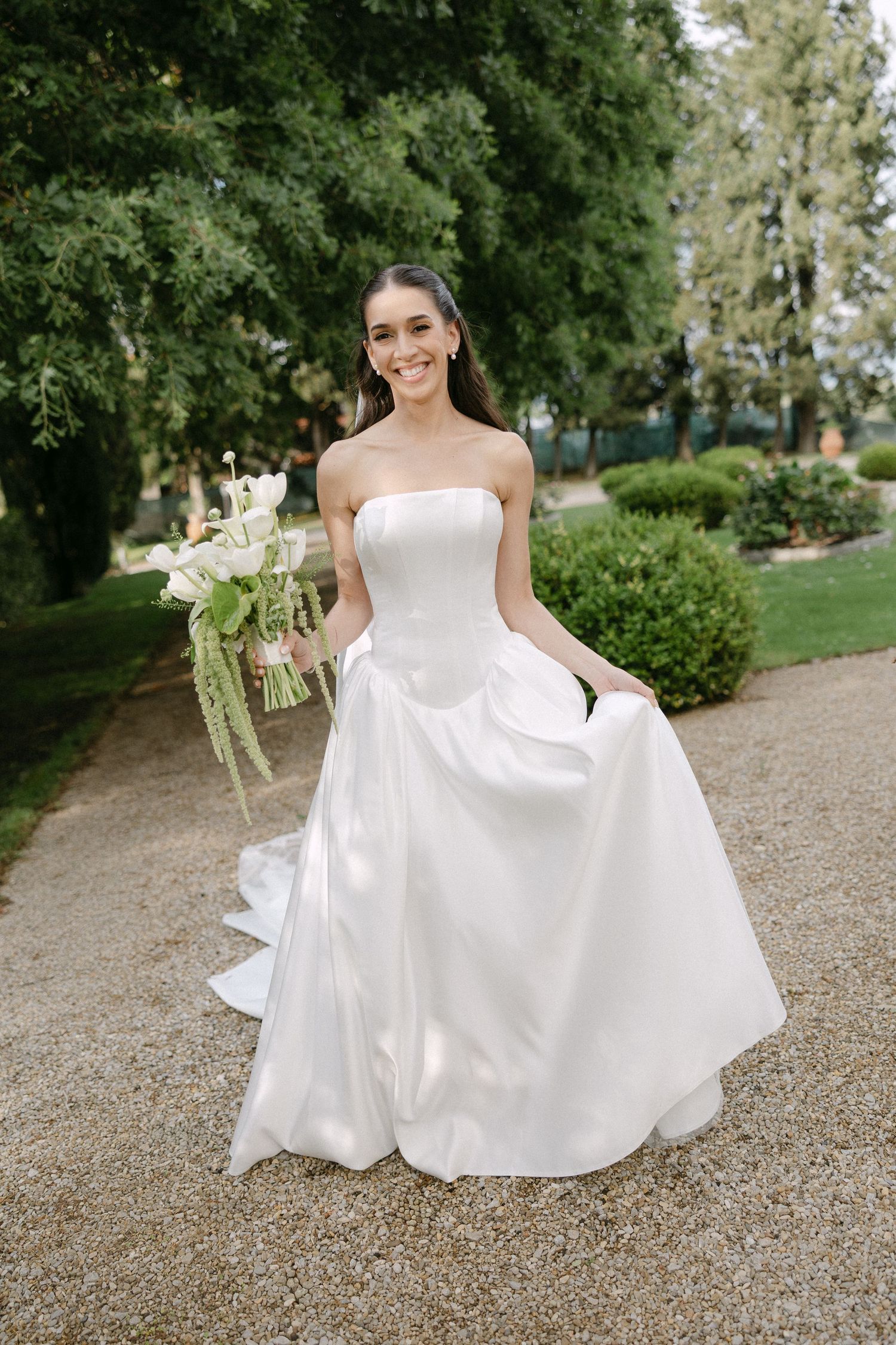 Smiling bride in strapless white ball gown holds green and white bouquet on garden path.