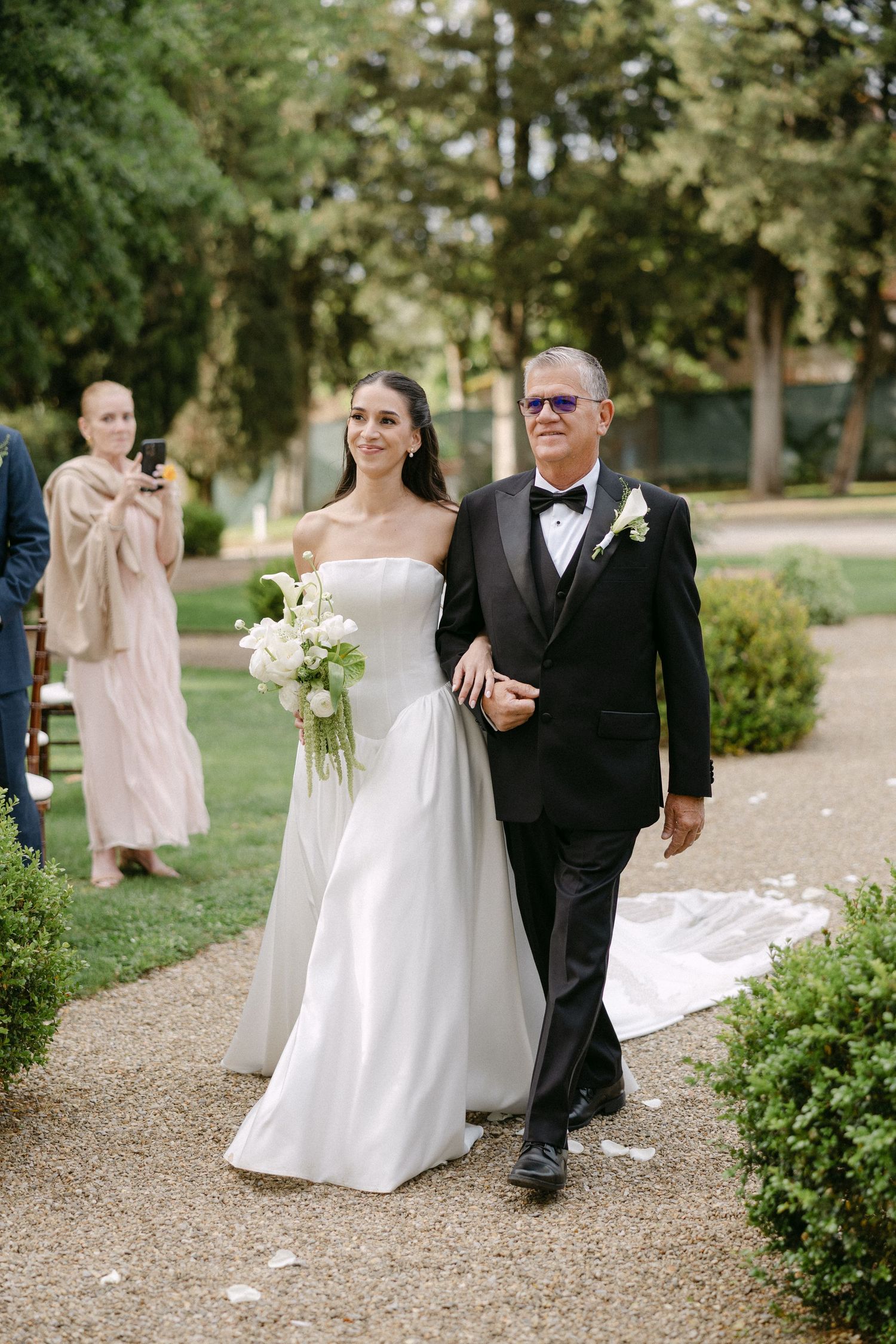 Bride walking down outdoor aisle escorted by father in tuxedo, holding white floral bouquet at garden wedding.