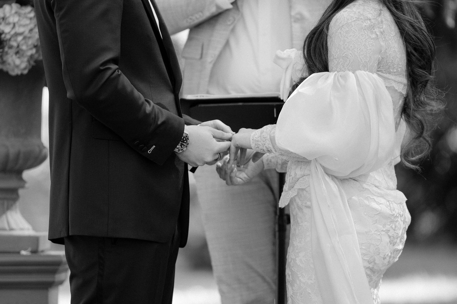 Black and white wedding photo of bride placing ring on groom's finger during outdoor ceremony.