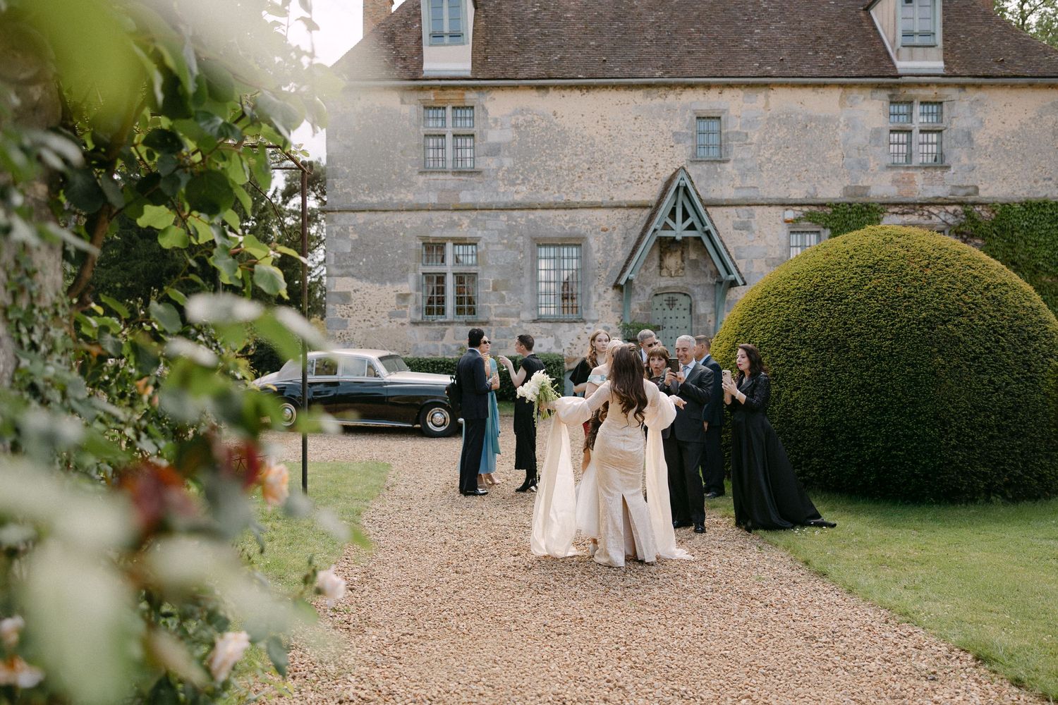 Wedding guests gathering outside a historic French manor house with vintage car and topiary hedges.