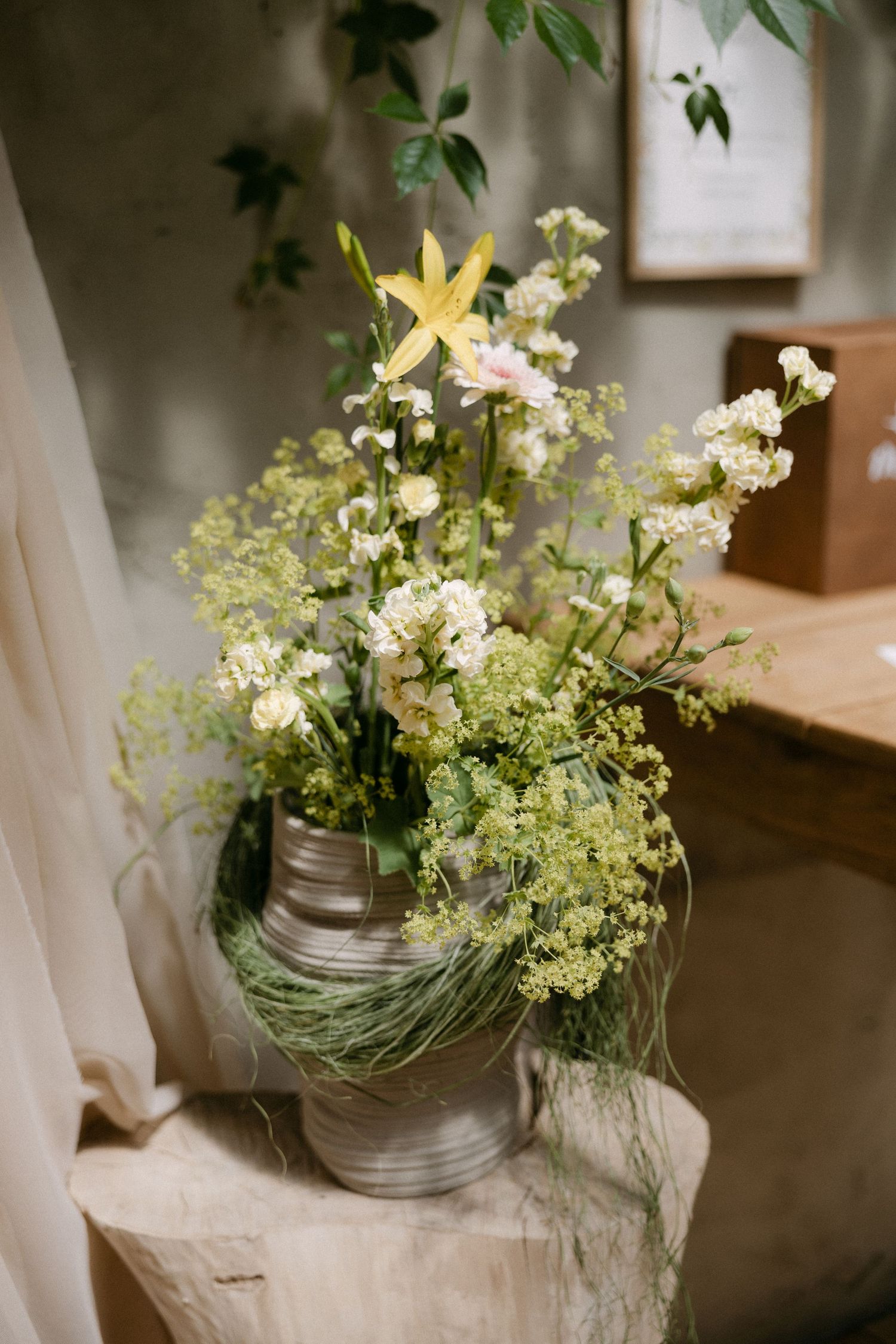 Rustic floral arrangement with white blooms, yellow lily, and greenery in a silver bucket vase on a wooden stump.