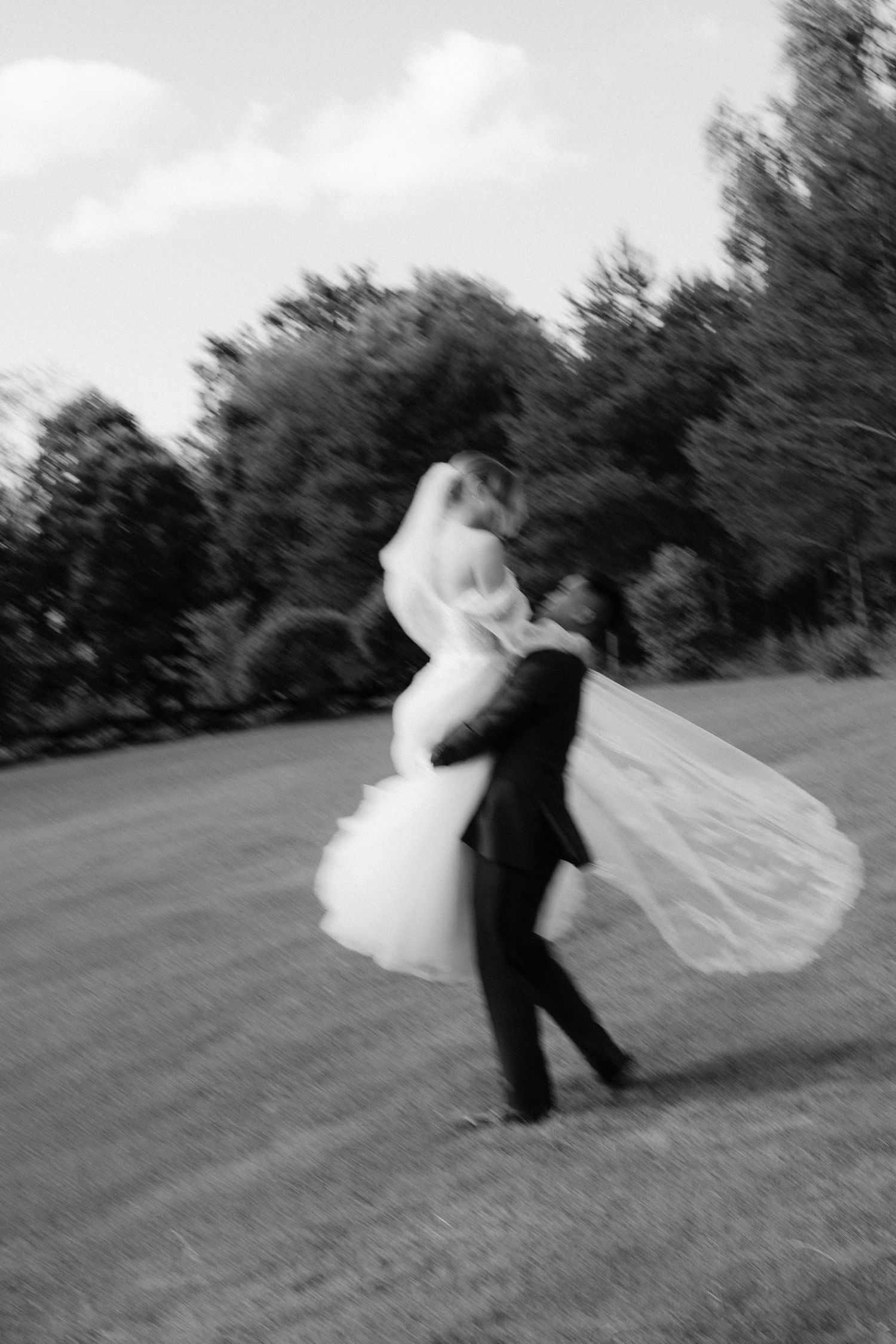 Black and white photo of groom spinning bride in wedding dress outdoors with motion blur effect.