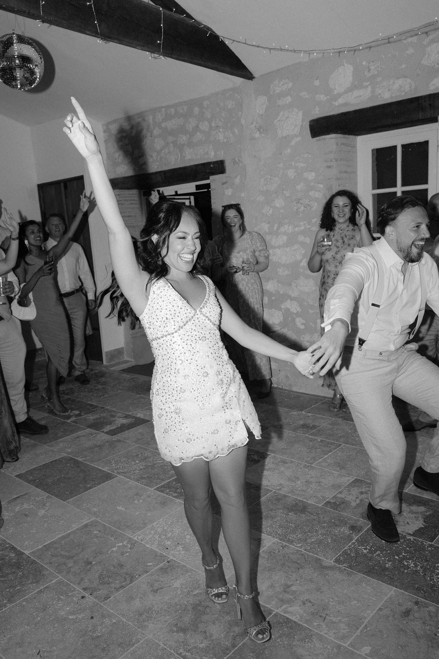Bride in sparkly mini dress dances joyfully with groom at lively black and white wedding reception.