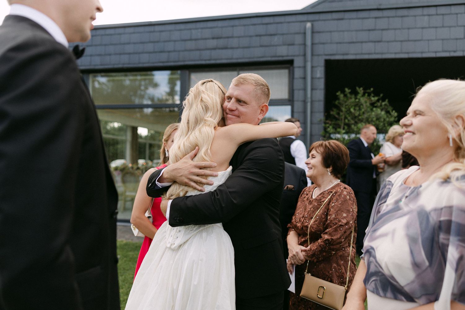 Bride in white dress receives warm hug from guest at outdoor wedding reception near modern building.