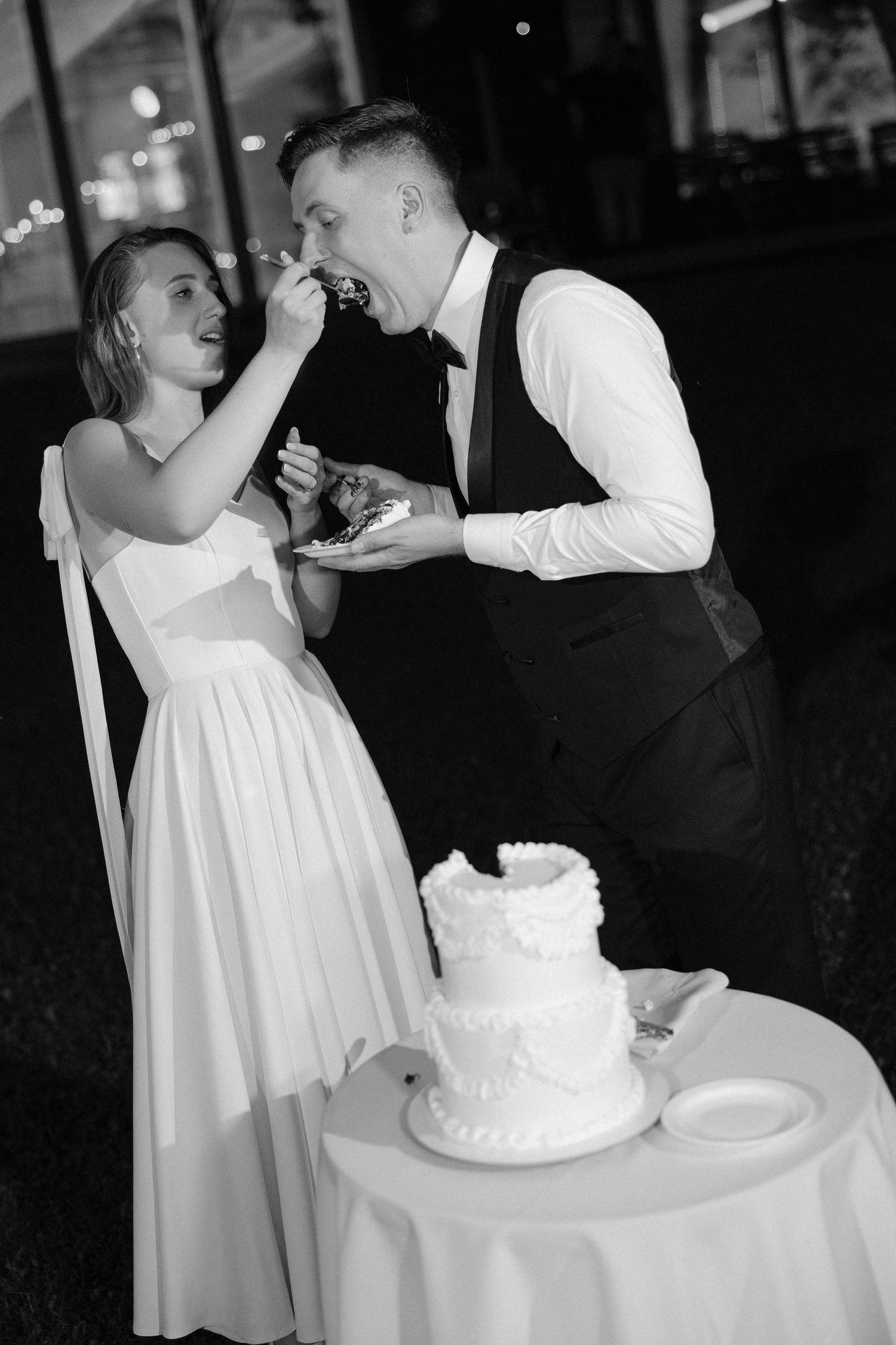 Bride and groom sharing wedding cake at reception in black and white photo with two-tier cake on table beside them.