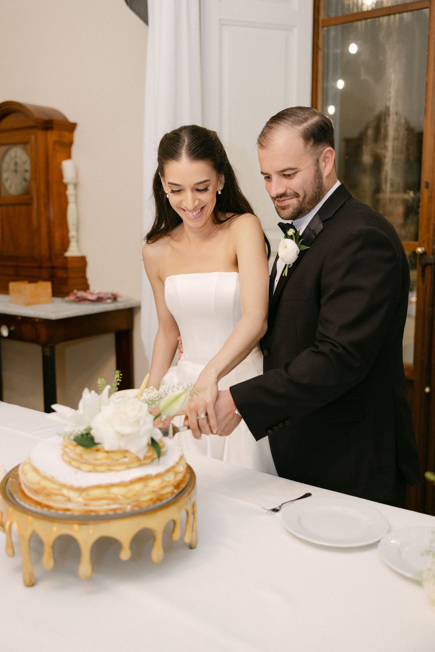 Smiling bride and groom cut elegant white floral-topped crepe wedding cake at reception table.