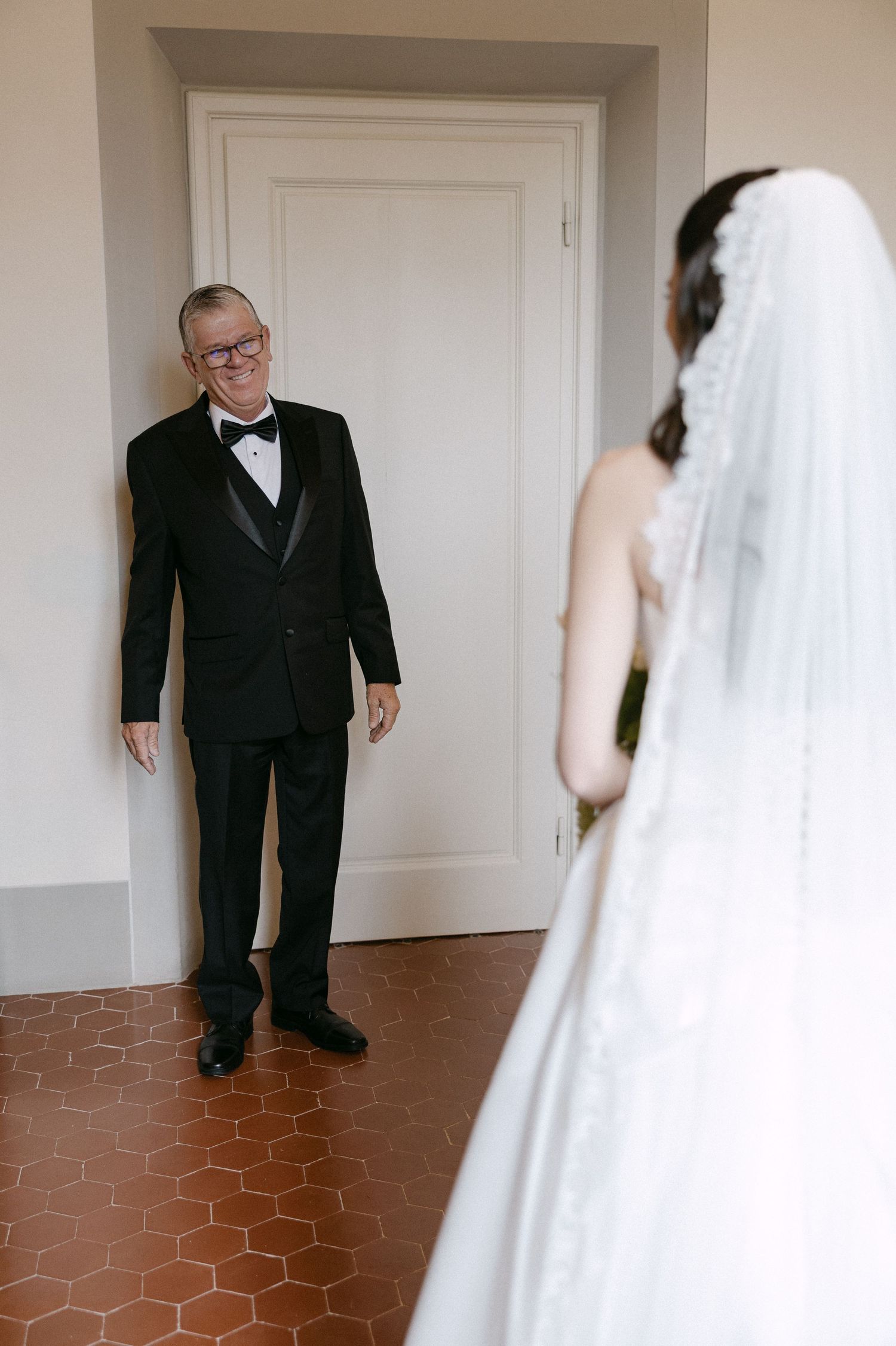 Father in tuxedo smiles seeing bride in white wedding gown and veil for first look moment.